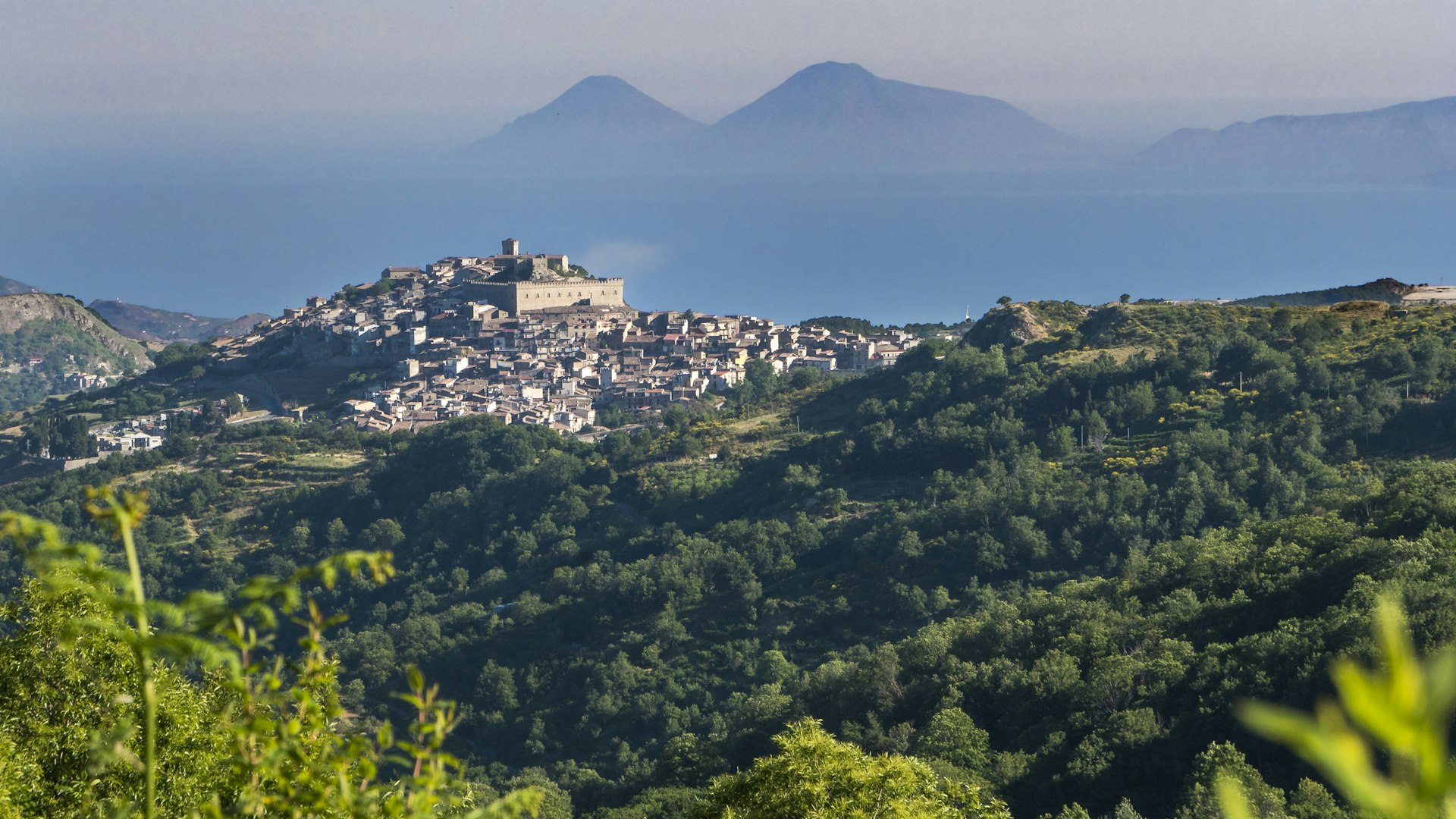 A medieval town on a hill surrounded by woodland with distant mountain peaks
