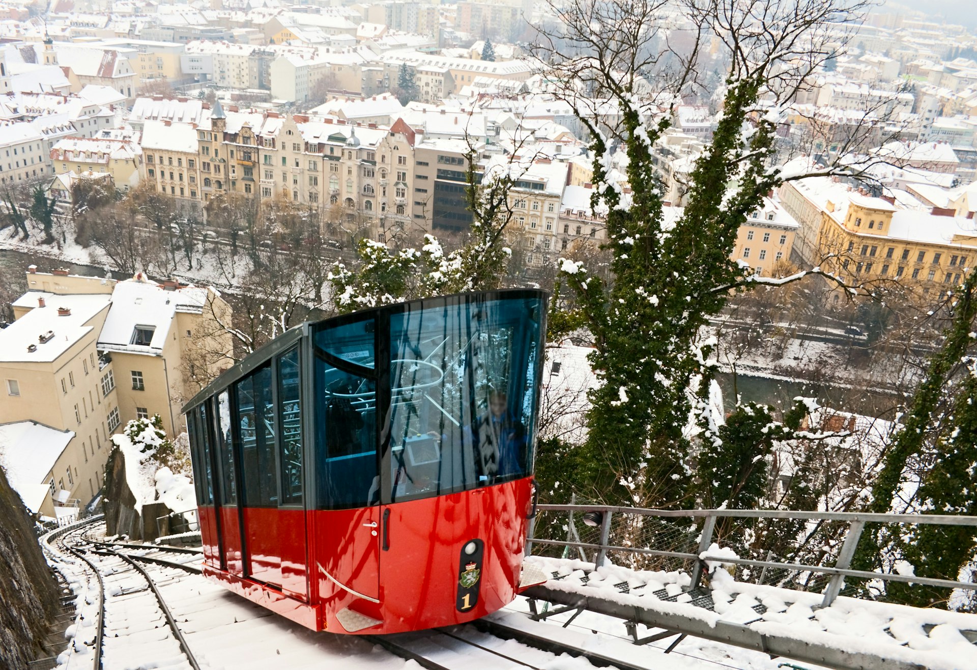 A red car on a funicular railway climbs up a steep, snowy track with the city of Graz, Austria, in the background.