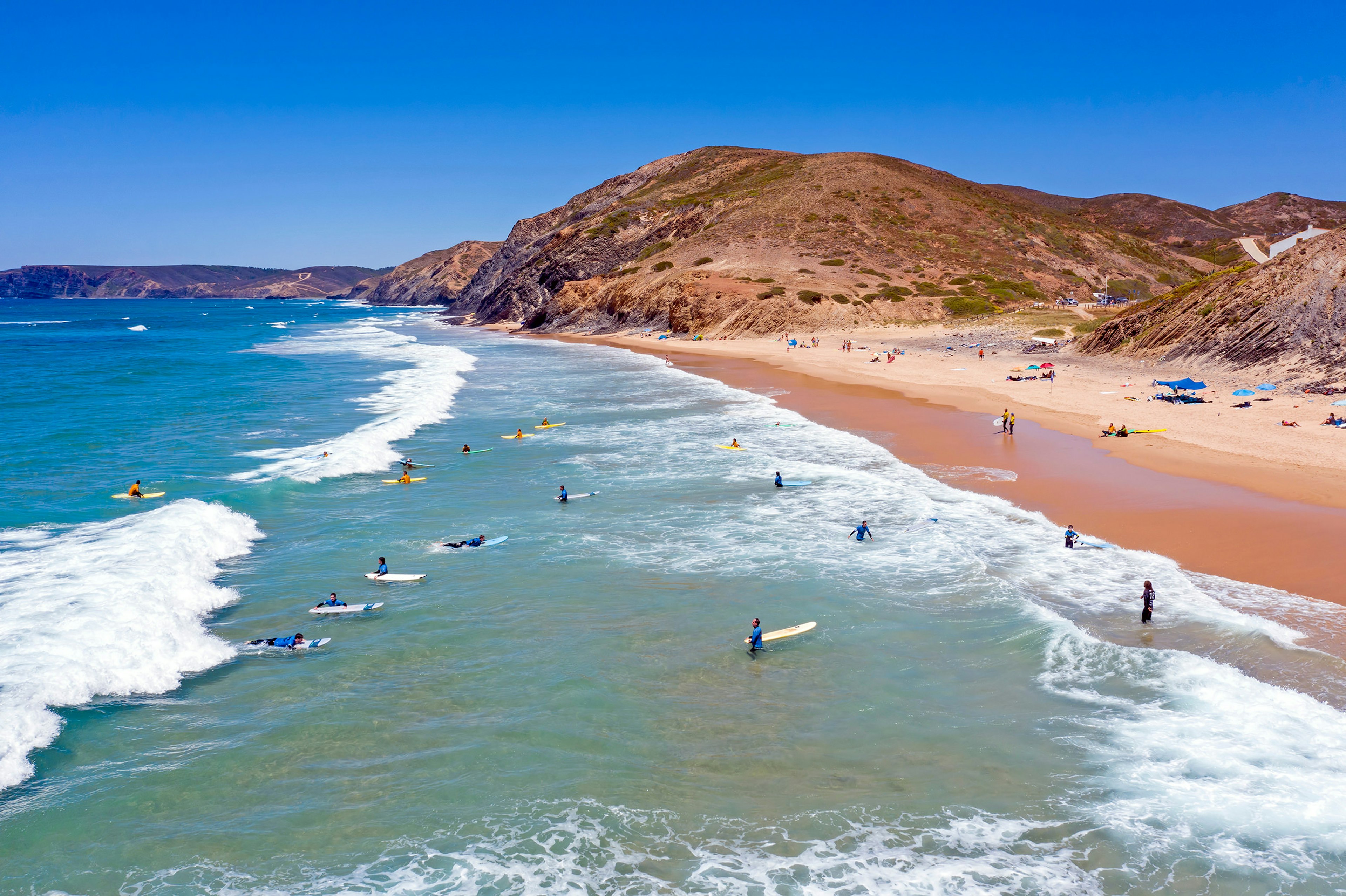 An aerial image shows surfers in the waters off a rocky red coast in Portugal.