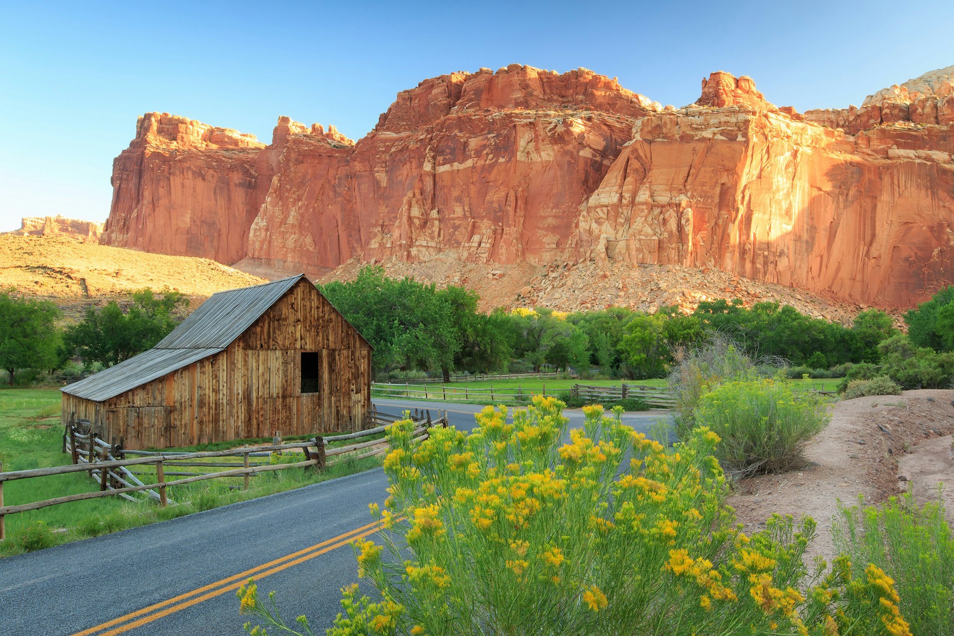 A historic barn sits at the edge of a wildflower meadow beneath towering red cliffs