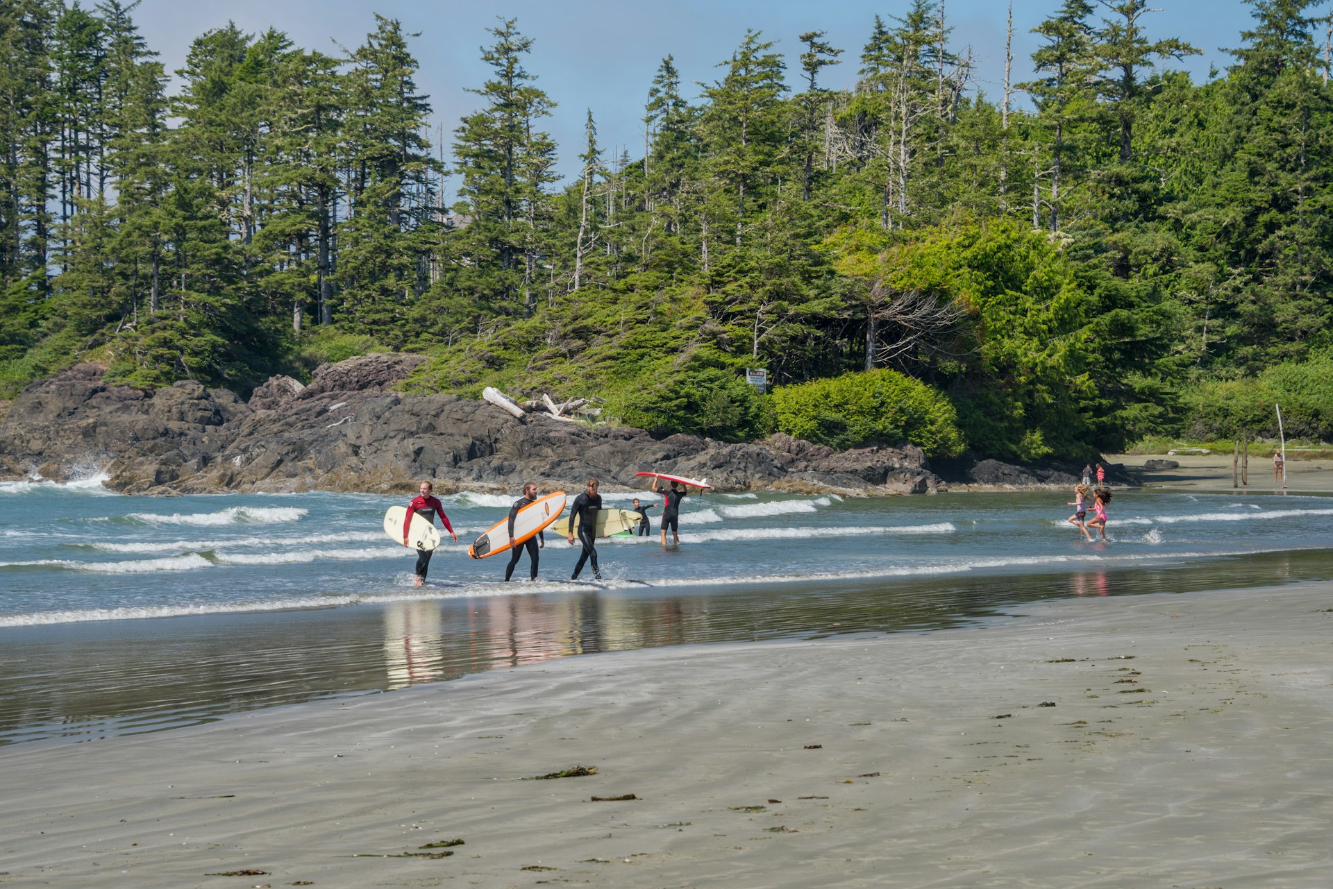 A small group of surfers are carrying surfboards as they leave the water and walk onto the sand of Long Beach, Vancouver Island