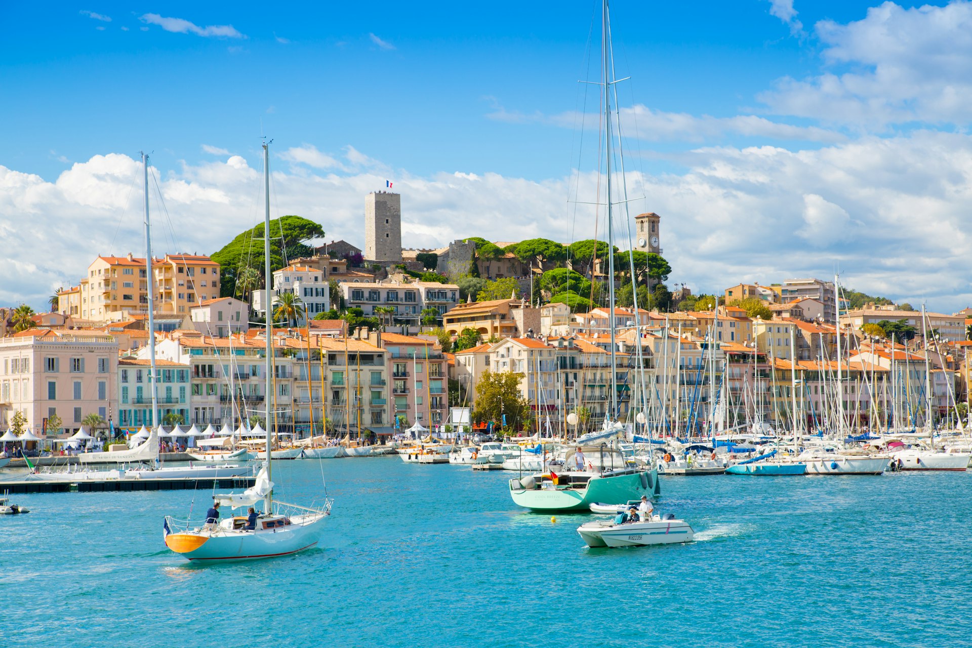 Many small sailing boats are moored in the Port of Cannes, with traditional French houses built up the hill behind