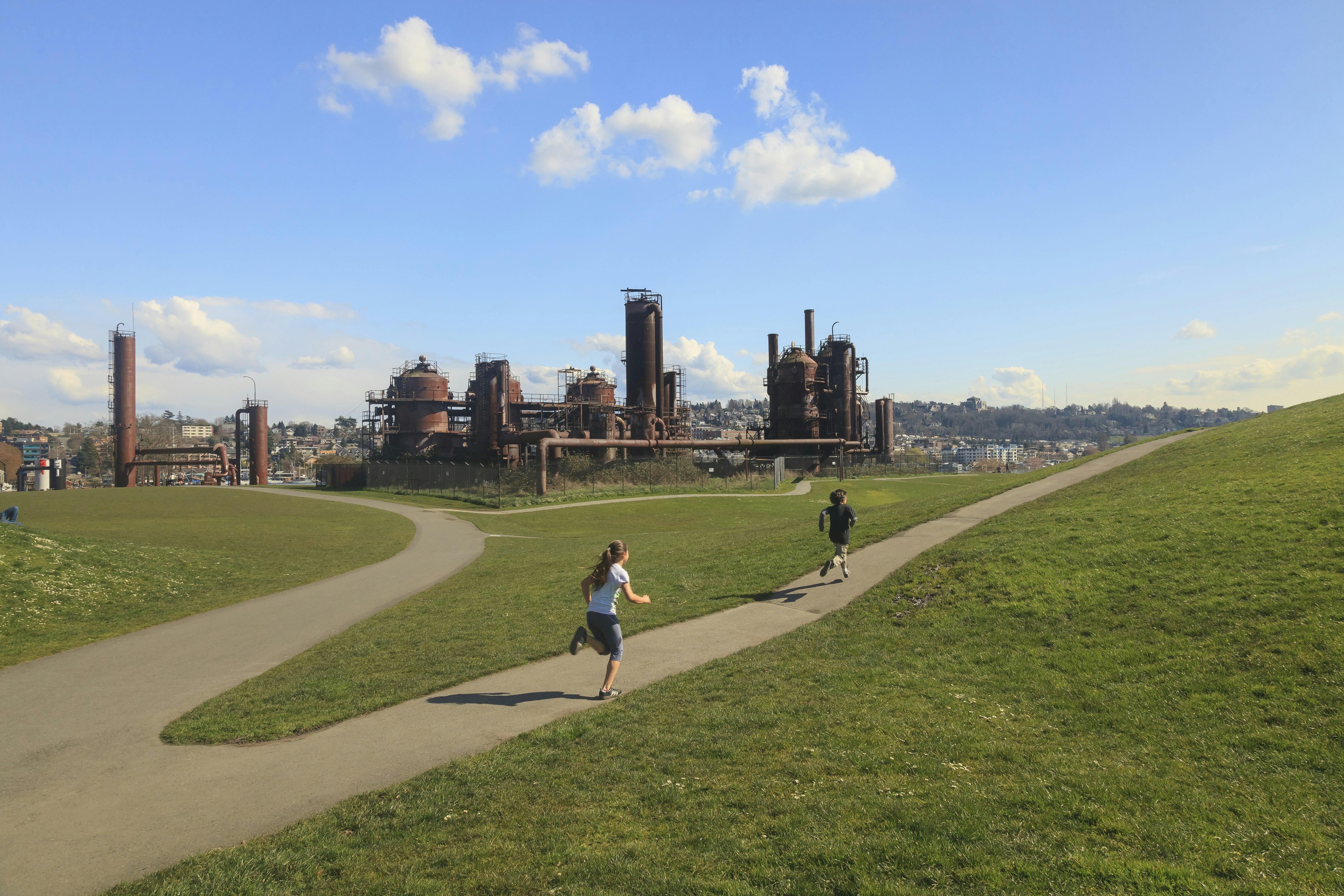 Two young children run along a path in a park on a sunny day with the rusty hulk of old gas works in front of them