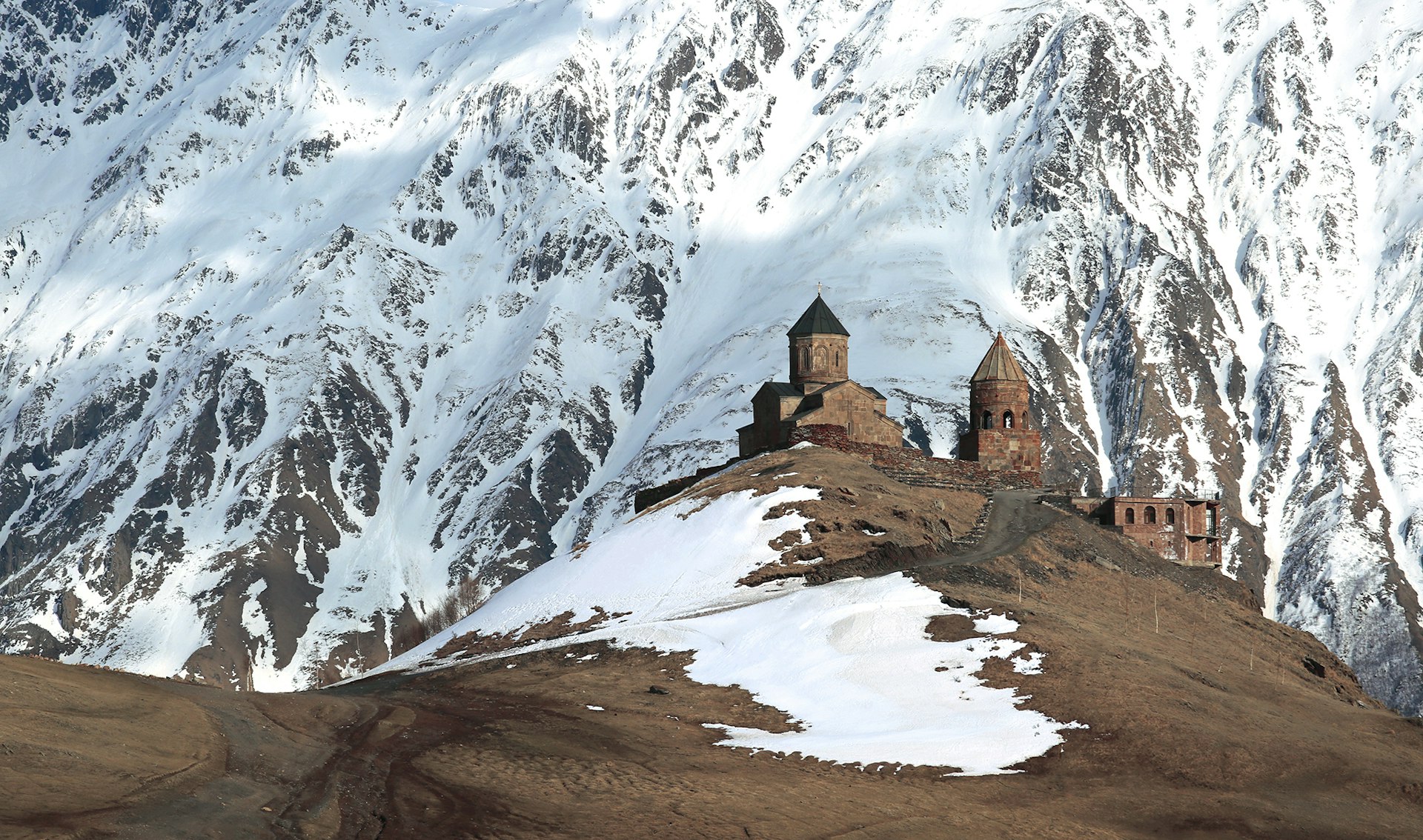 A monastery in Georgia sits atop a hill, with incredible snowy mountains in the background. 