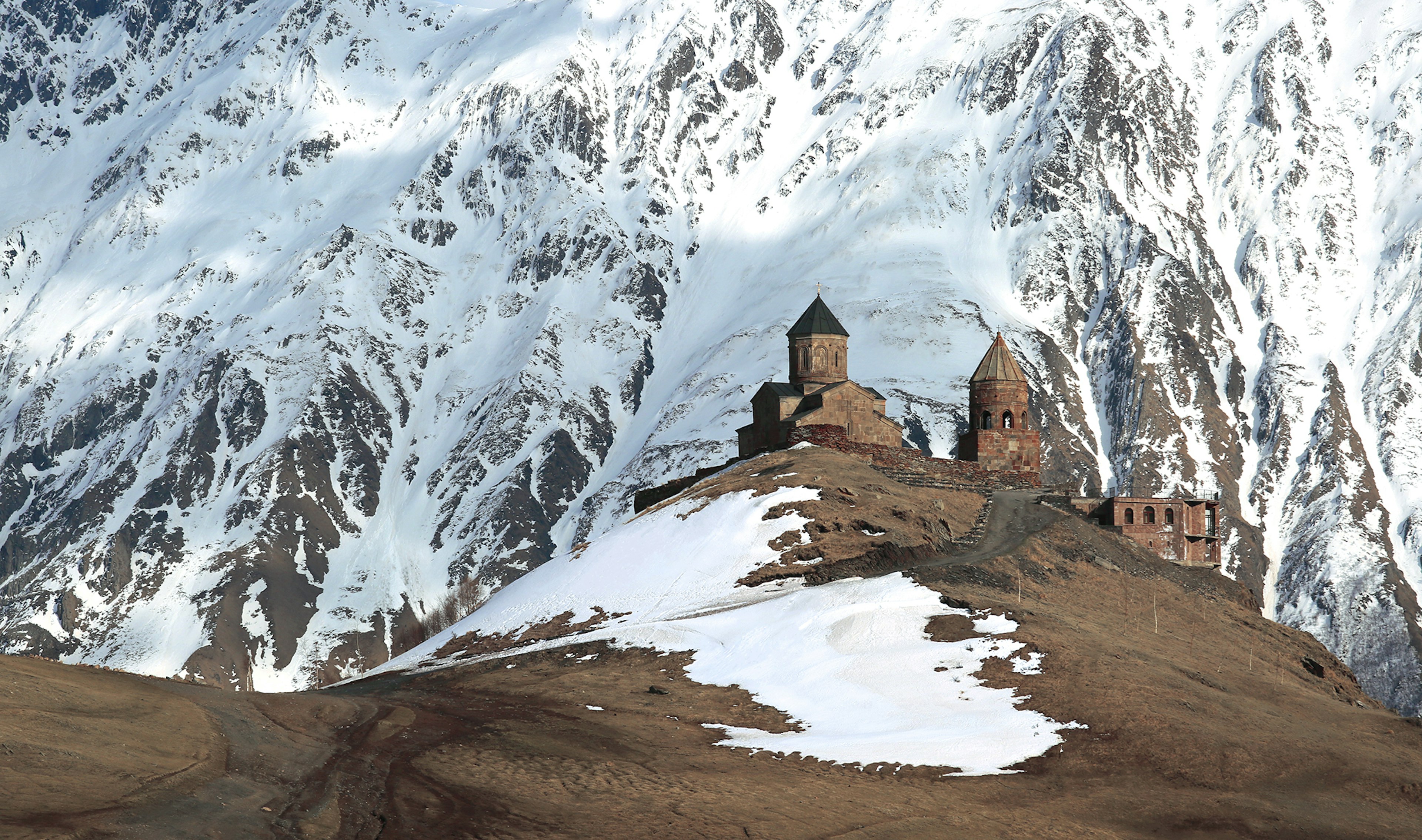 A monastery in Georgia sits atop a hill, with incredible snowy mountains in the background.