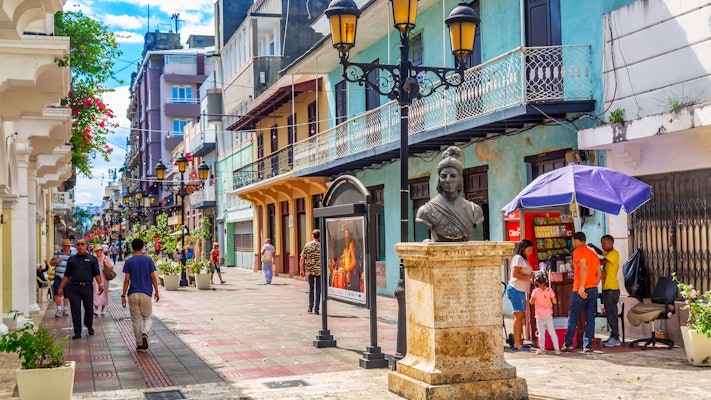 People walk along a pedestrian street past colorful buildings in the Dominican Republic.