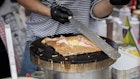 Hands of cook in gloves preparing crepe galette, typical of Brittany, France, with ham, cheese and egg on black griddle.