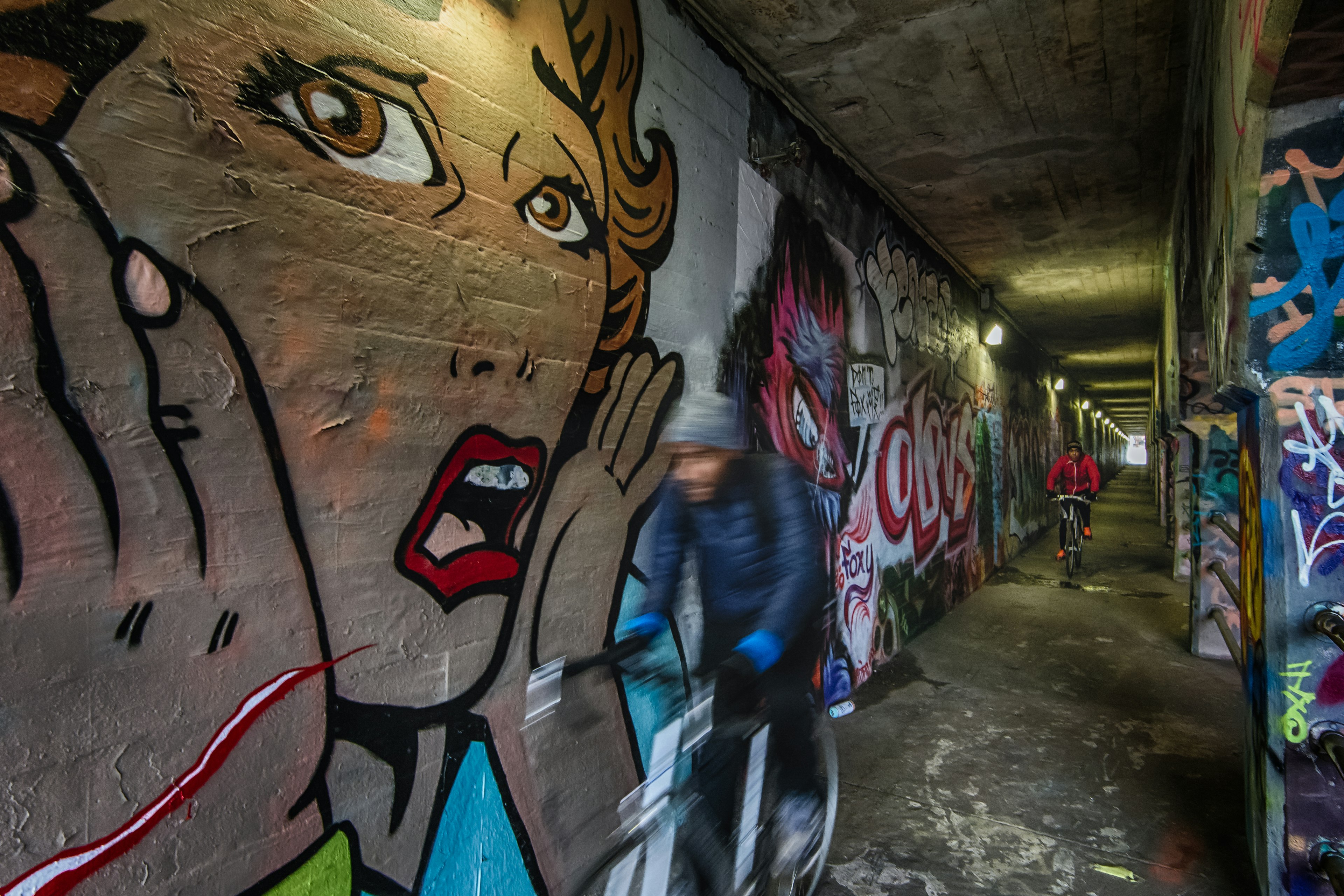 A large artwork of a shocked woman is painted in the style of Roy Lichtenstein on the wall of a graffiti-covered tunnel, while cyclists ride past