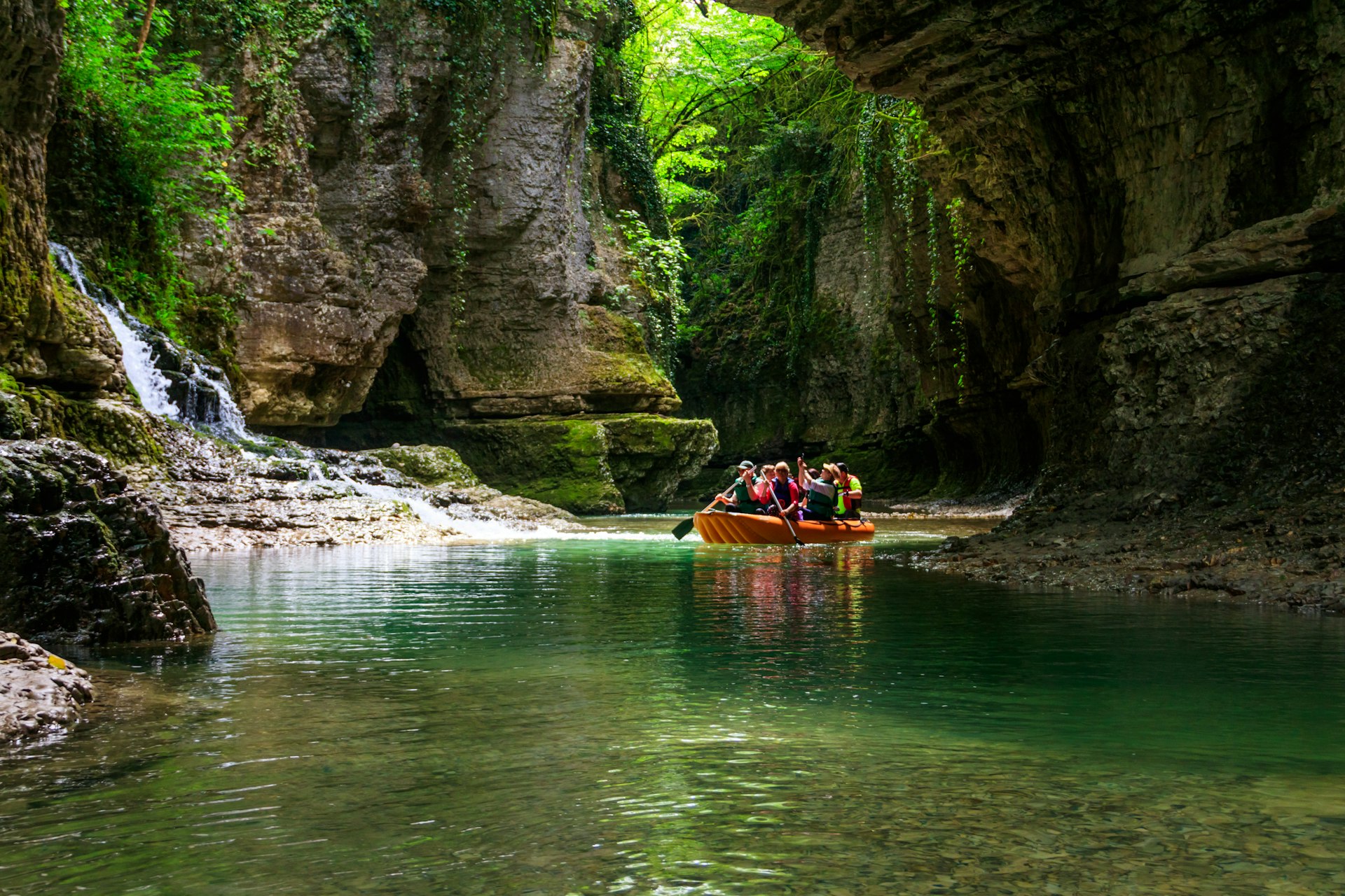 A group of people are paddling a raft along a blue-green river in a canyon