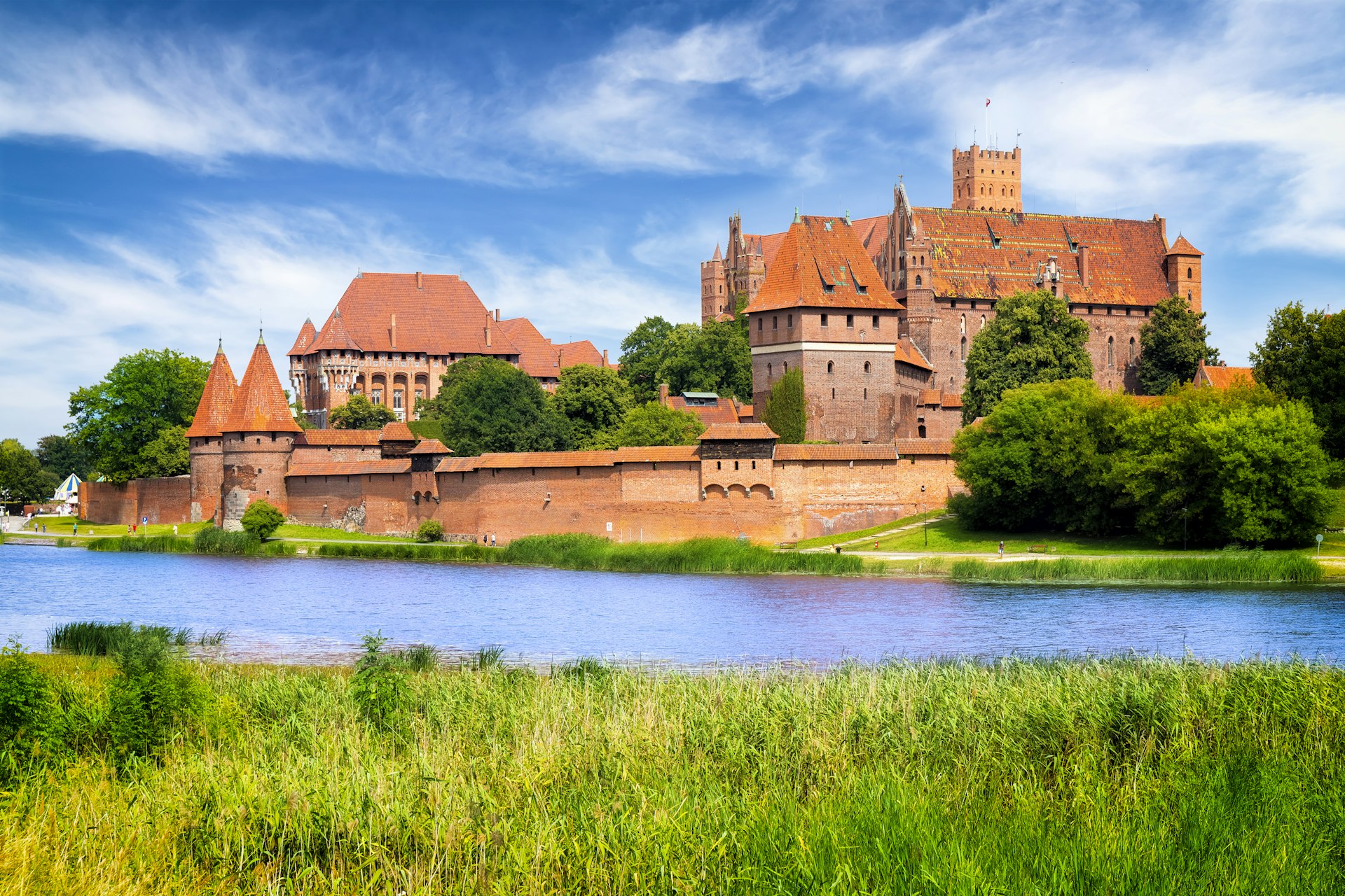 Um enorme castelo de tijolos vermelhos com muitas torres e ameias fica às margens de um rio calmo.
