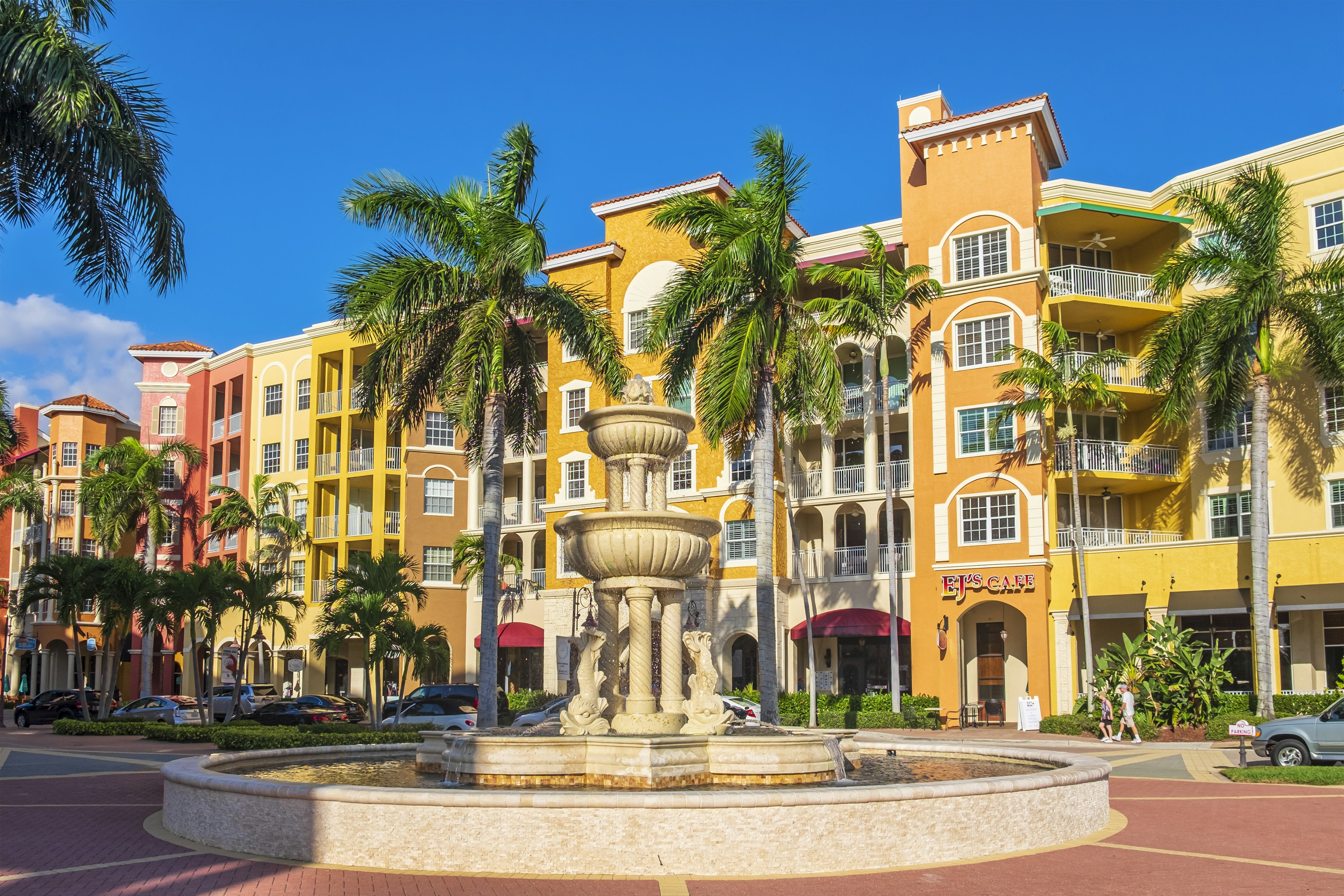 A fountain with palm trees stands in front of a row of tall, colorful, European-style buildings