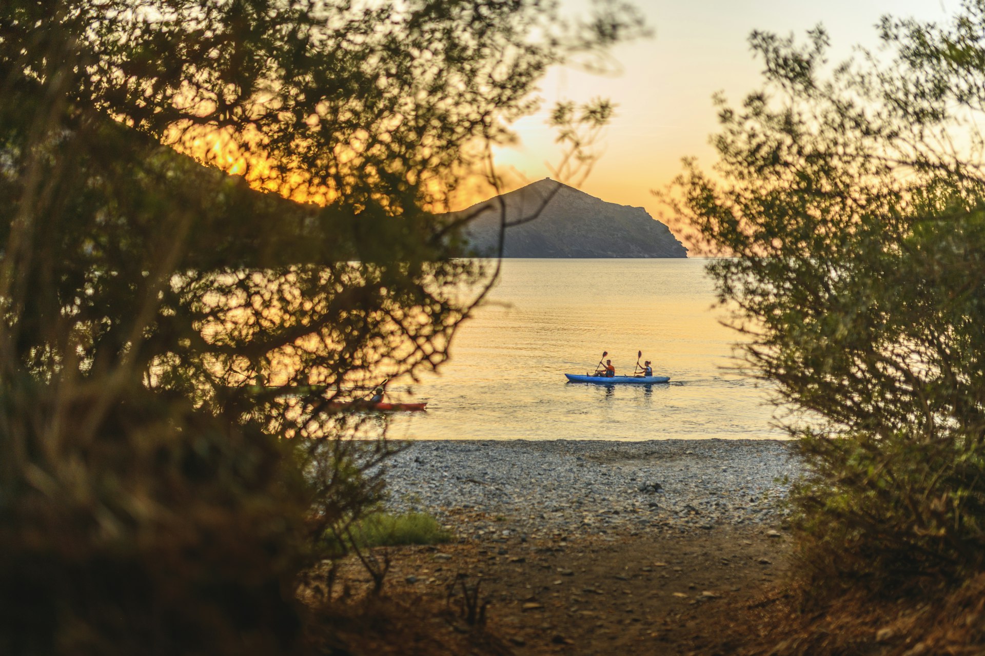 Distant perspective of friends paddling kayak along shore of Mediterranean Sea with sun rising over headland in background.