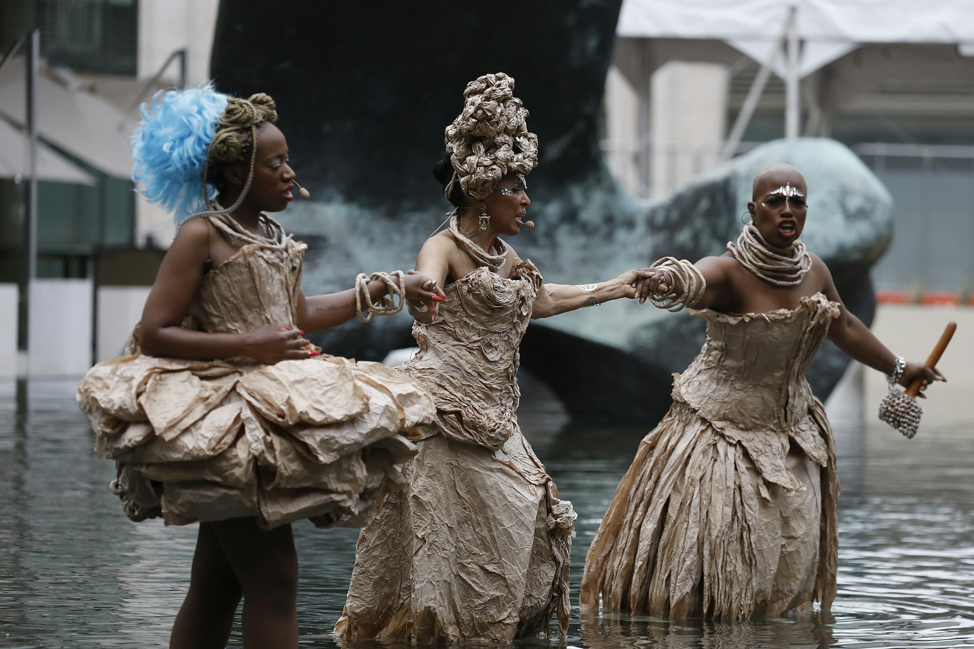 Three women in beige and blue costumes stand in the water during a performance outside Lincoln Center