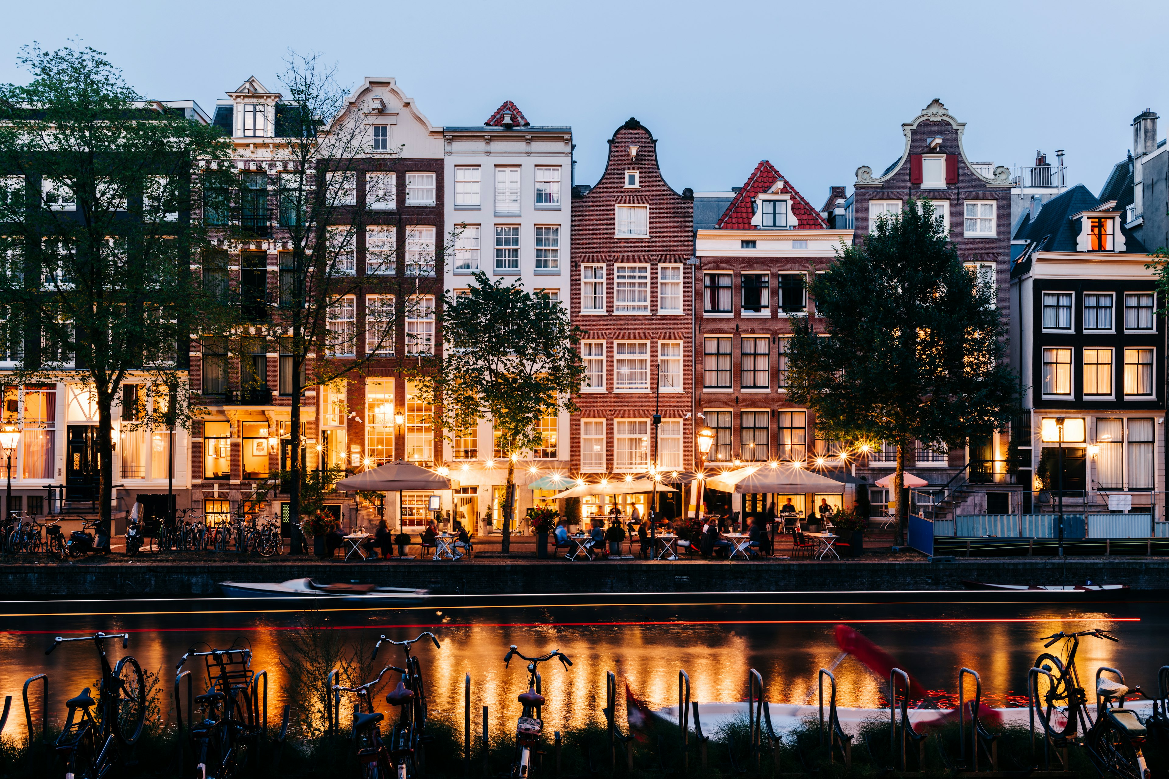 People sit at canalside tables outside bars and restaurants lit by string lights in the evening