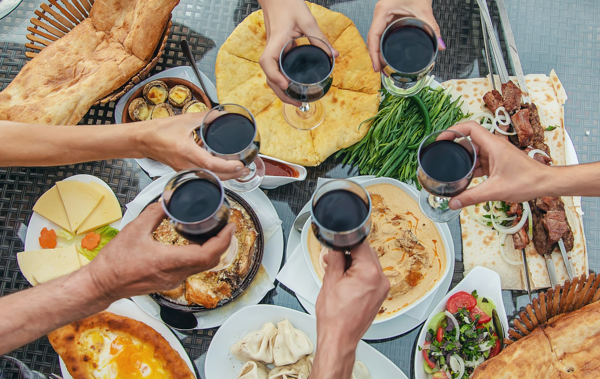 Six people raise a toast with glasses of red wine over a table laid with many different traditional Georgian dishes