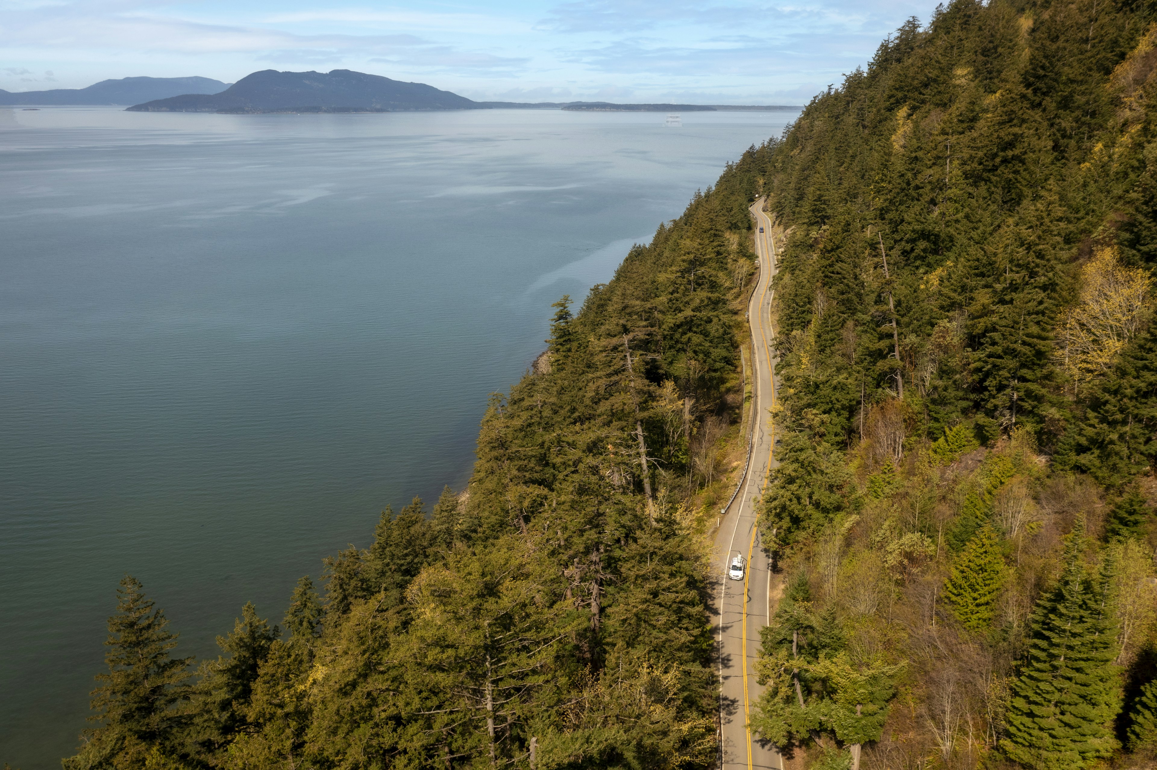 A car drives along a coastal highway, with the sea on one side and fall trees on the other.