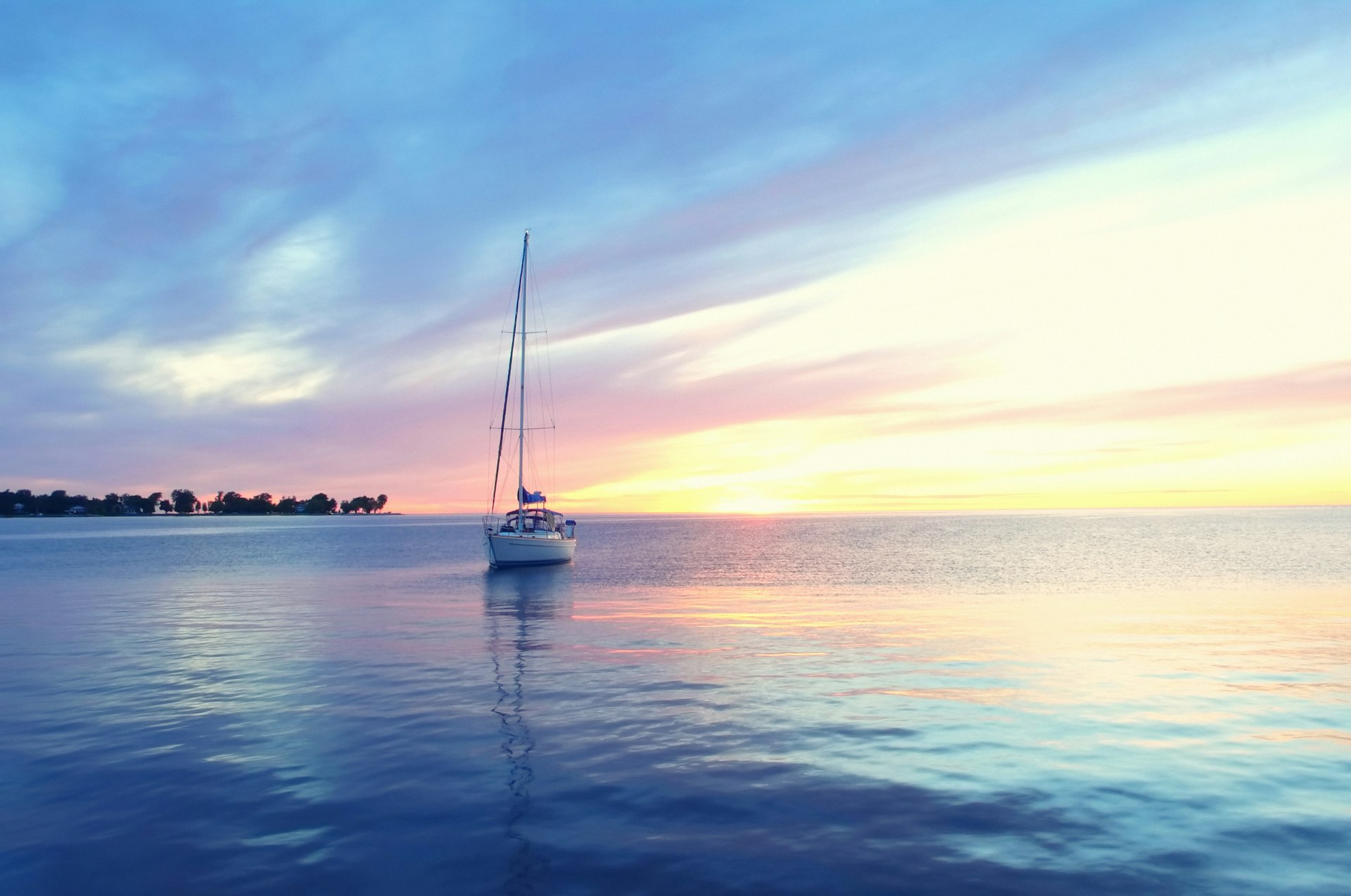 A single sail boat anchored in calm waters as the sunset casts pastel colors across the sky