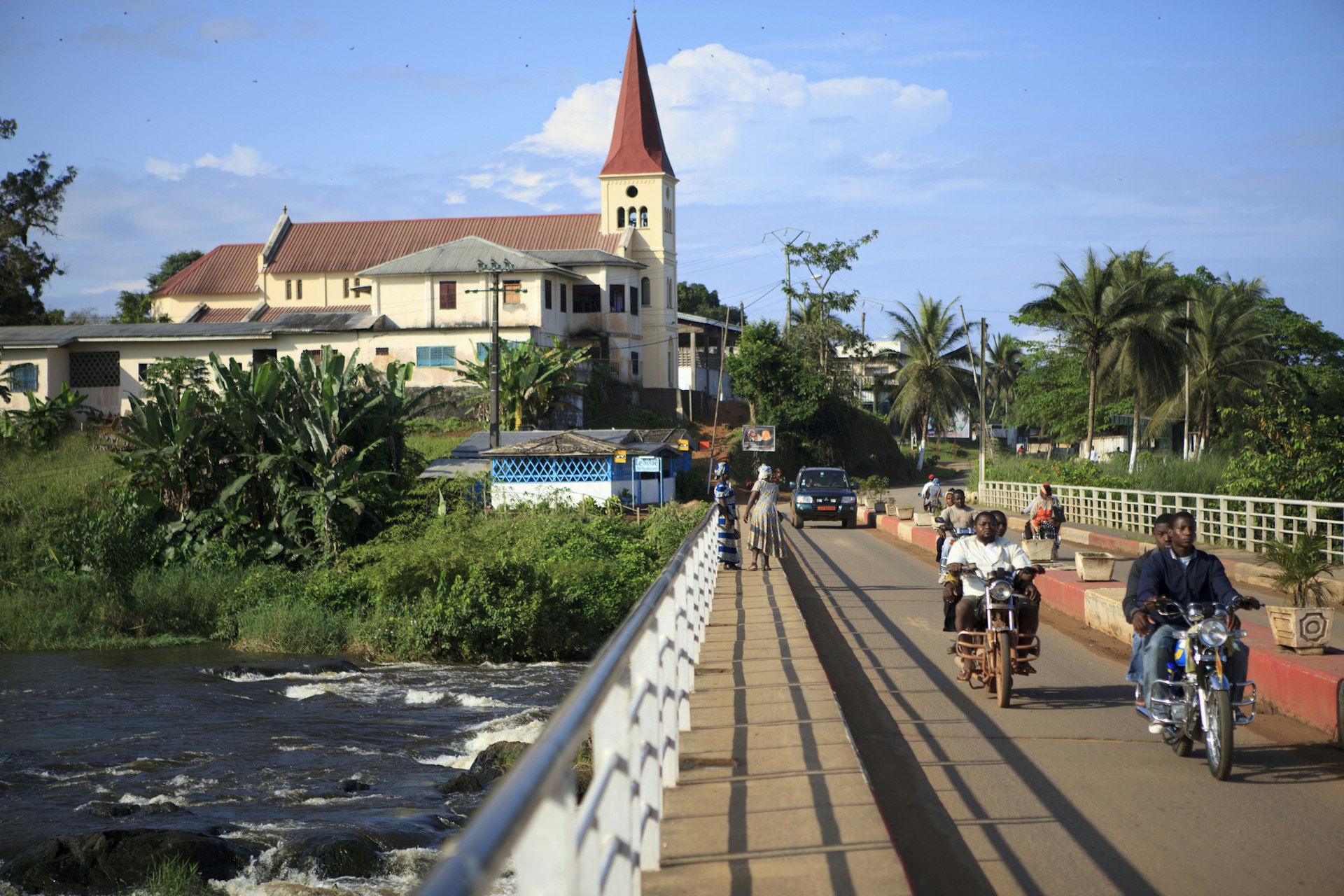 Motorcyclists cross a bridge in front of a church in a small town