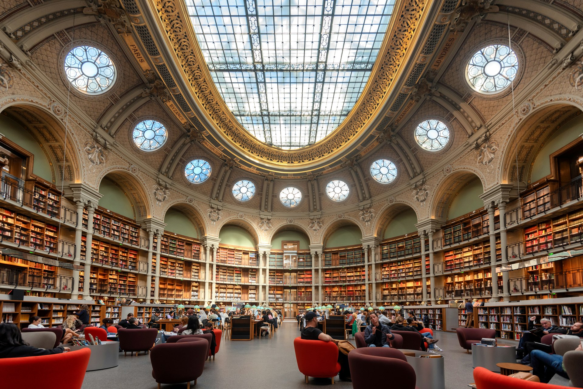 Many people are studying at long desks or lounging in colorful armchairs in a library. Many thousands of books are stored in shelves lining the walls and there is an enormous decorative oval skylight in the ceiling,