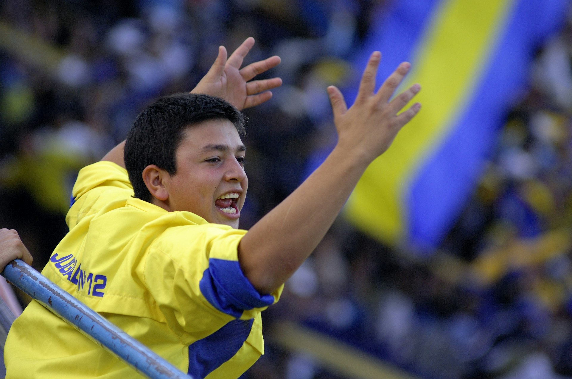 A teenager wearing the yellow and blue colors of the Boca Juniors team is raising his arms and cheering at a soccer match in Buenos Aires, Argentina