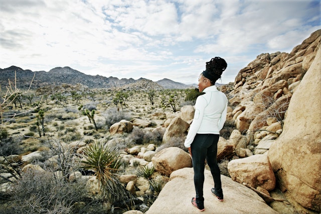 A woman in exercise gear stands on a high rock looking out over a desert landscape.