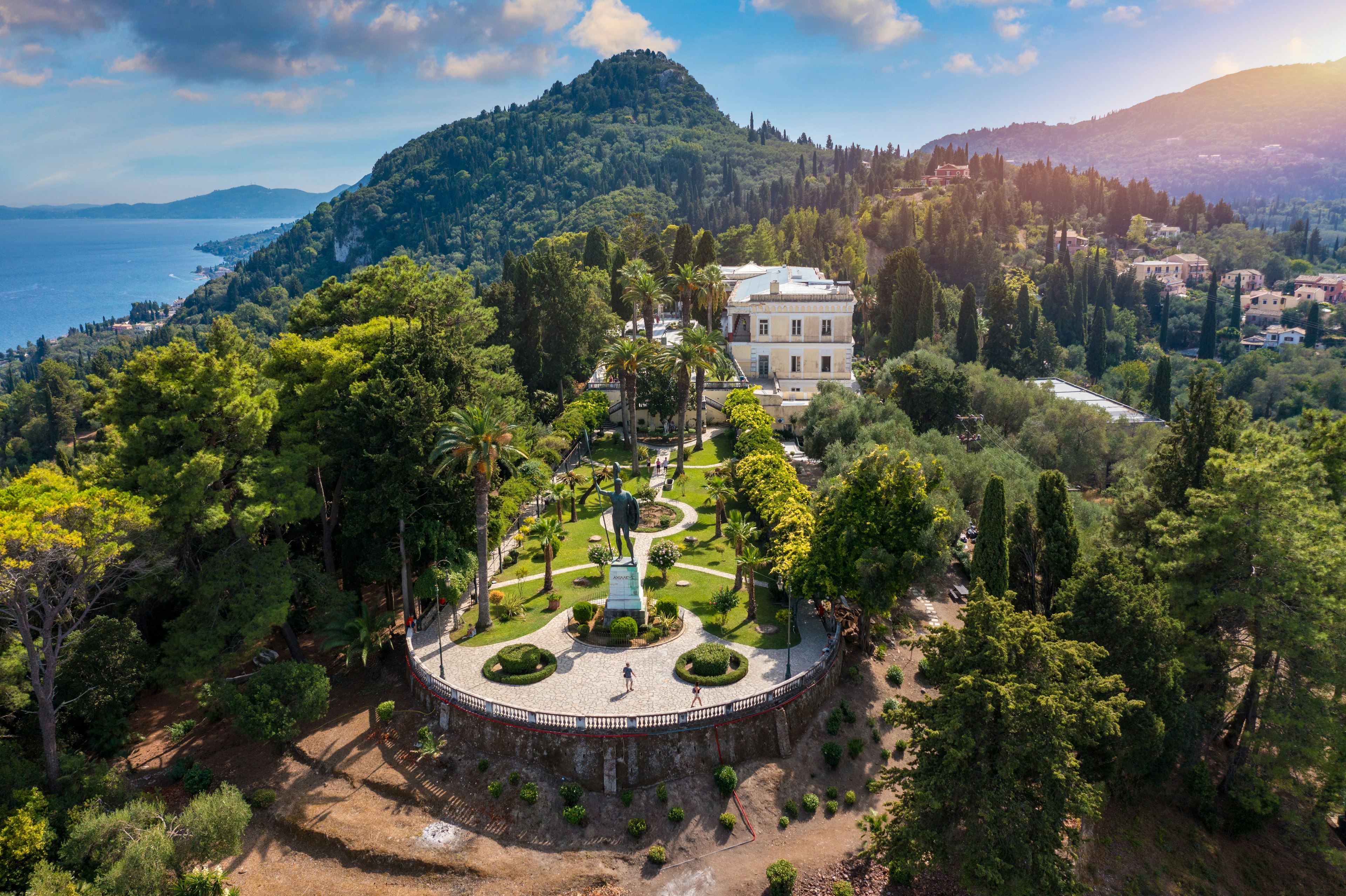 A white mansion-like building stands in the center of landscaped gardens with statues