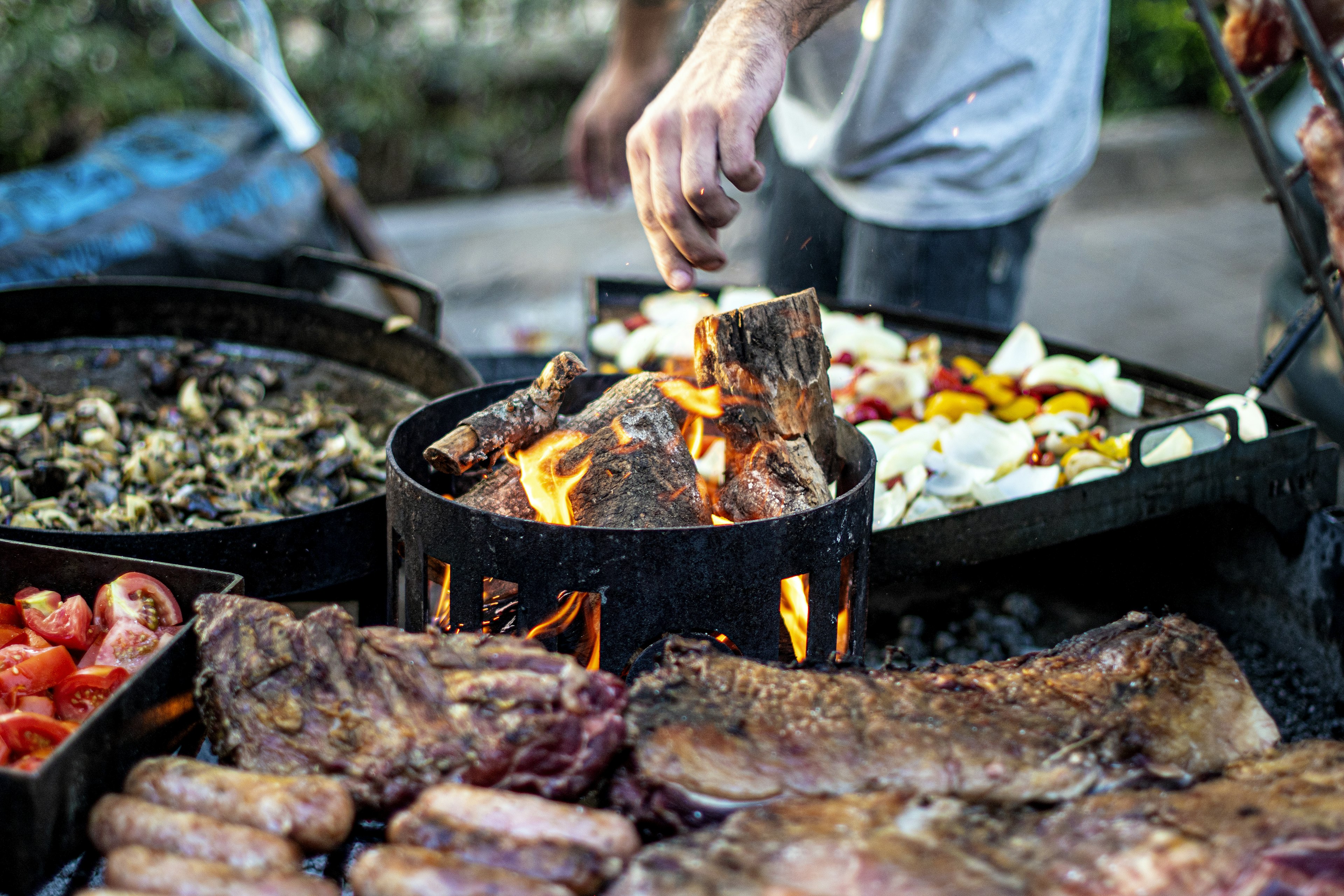 A man is tending to a large, wood-fired barbecue grill is loaded with various meats and vegetables