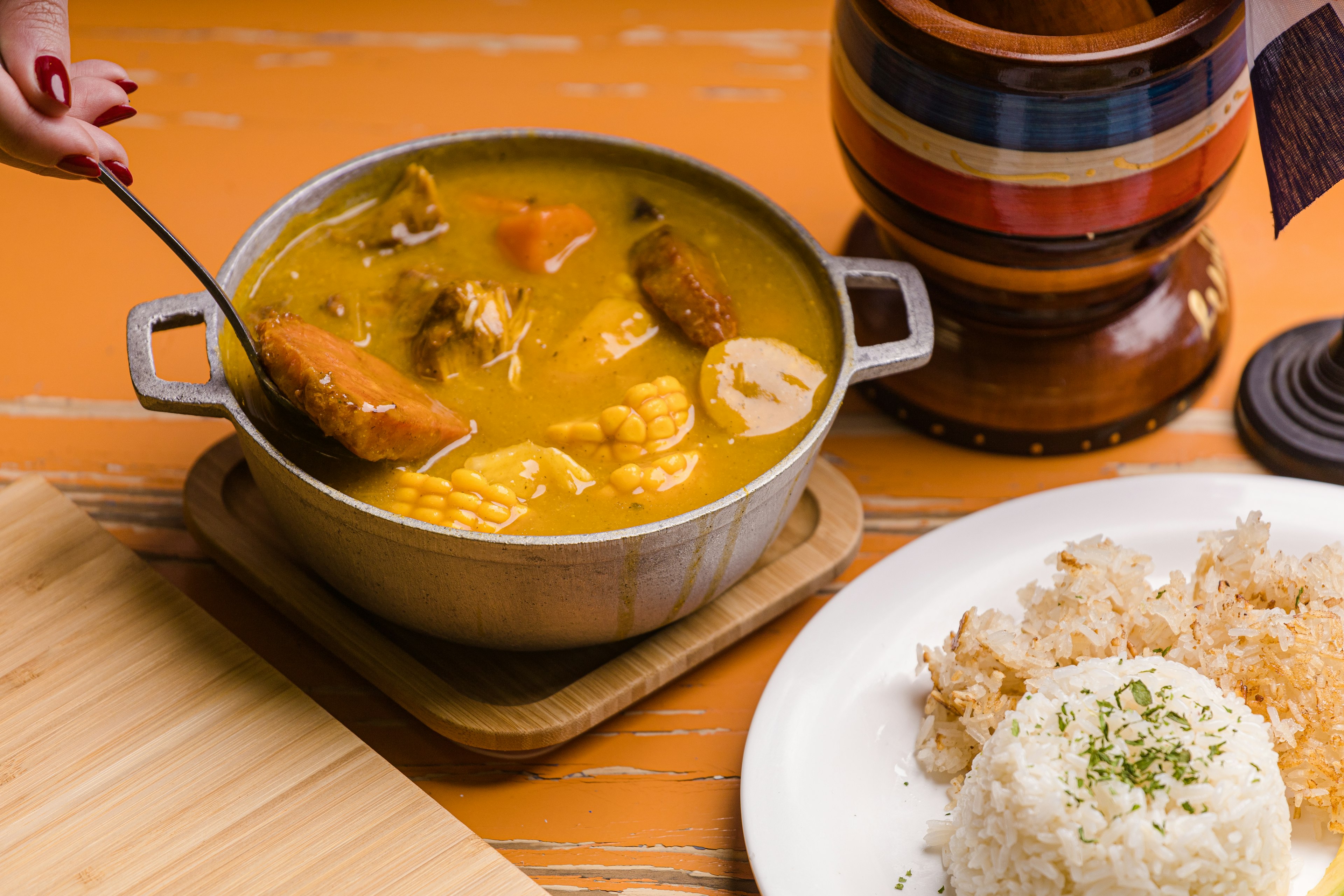 A bowl of meat stew with large pieces of corn, served with rice