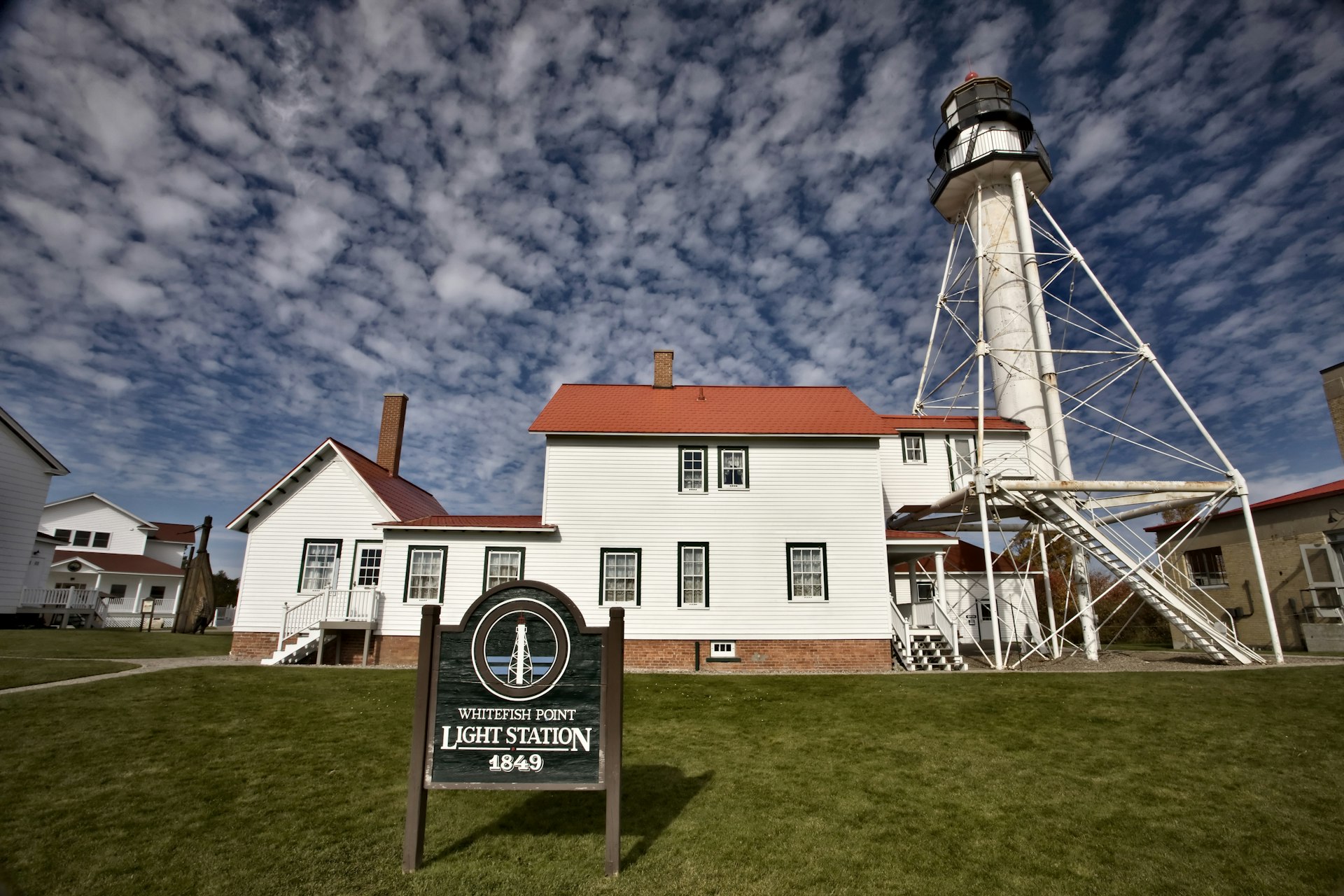 A lighhouse and red-roofed buildings. A sign reads "Whitefish Point Light Station 1849"