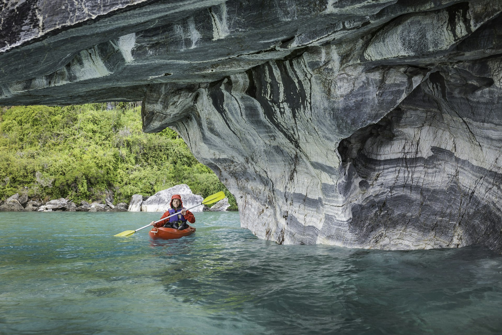 A kayaker leaves turquoise waters and enters a cave with grey and white streaked rocks
