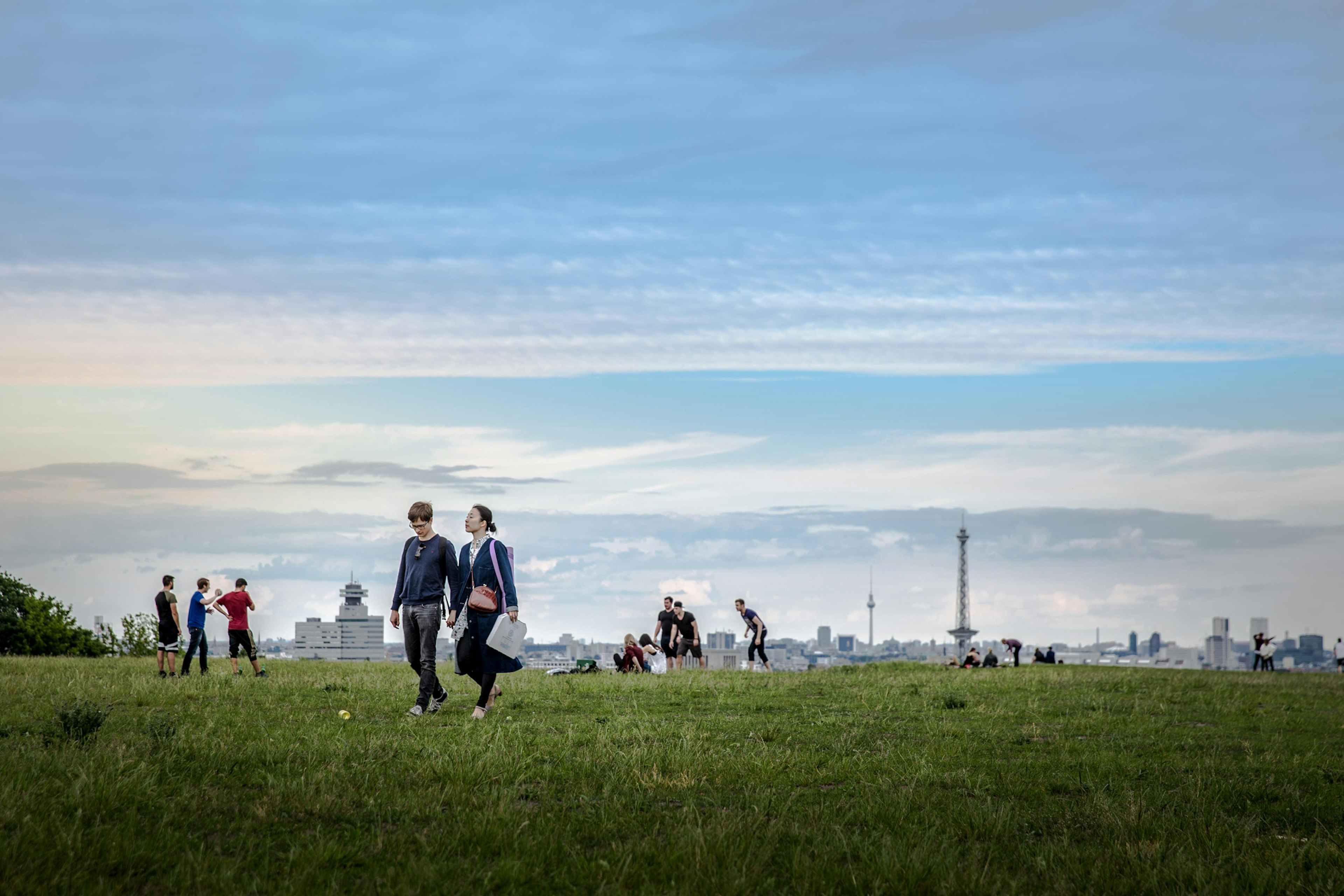 Two people walk hand in hand through a park as others play a game behind them. The cityscape rises in the distance