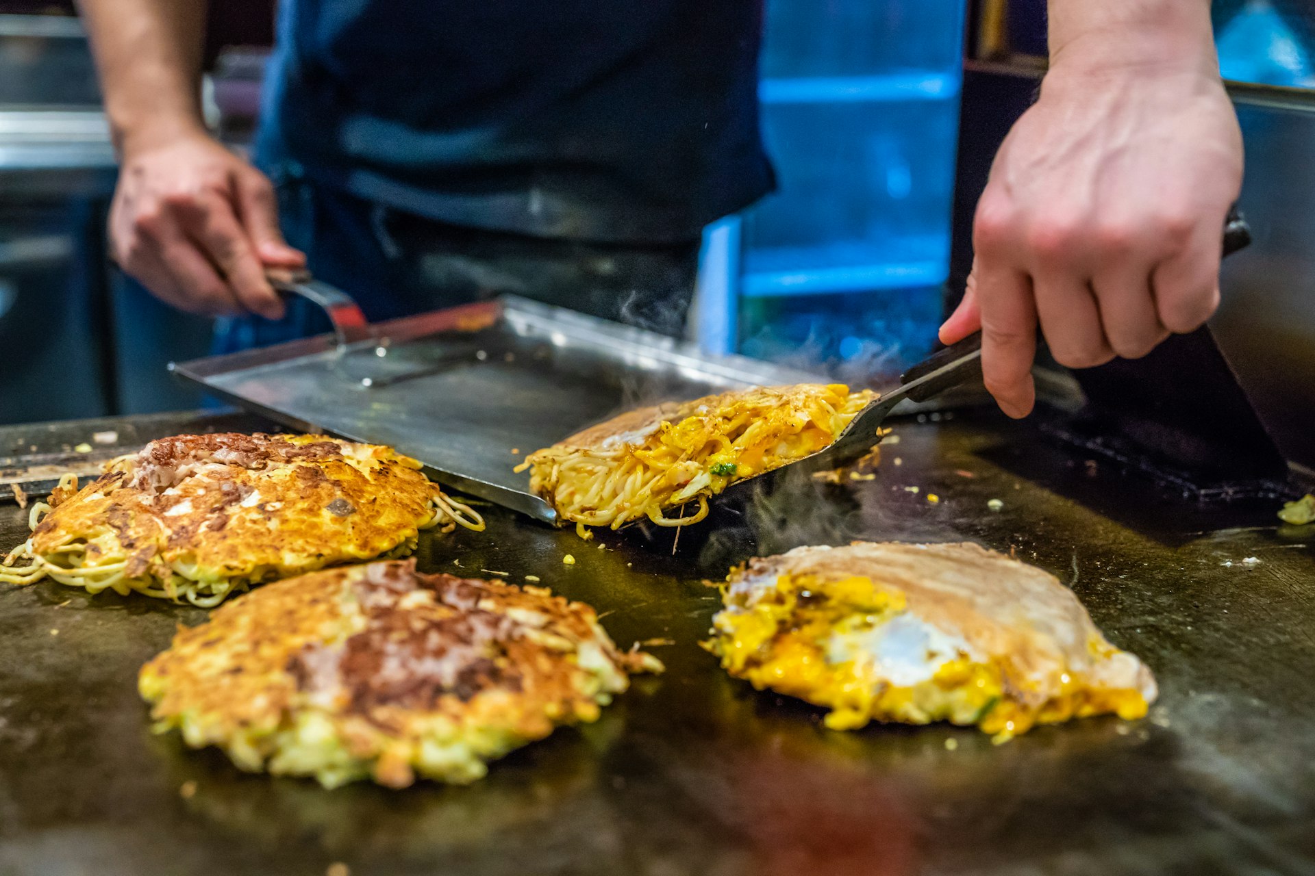 A cheg uses two flat spatulas to shift cabbage-based omelettes around on a hot plate