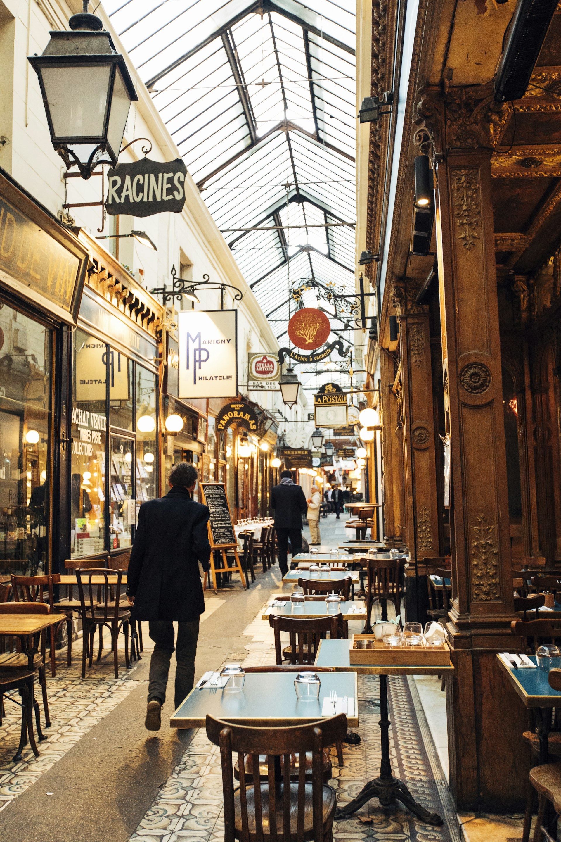 People walk through a narrow, glass-roofed passageway that is lined by restaurants and shops, some of which have dining tables outside.