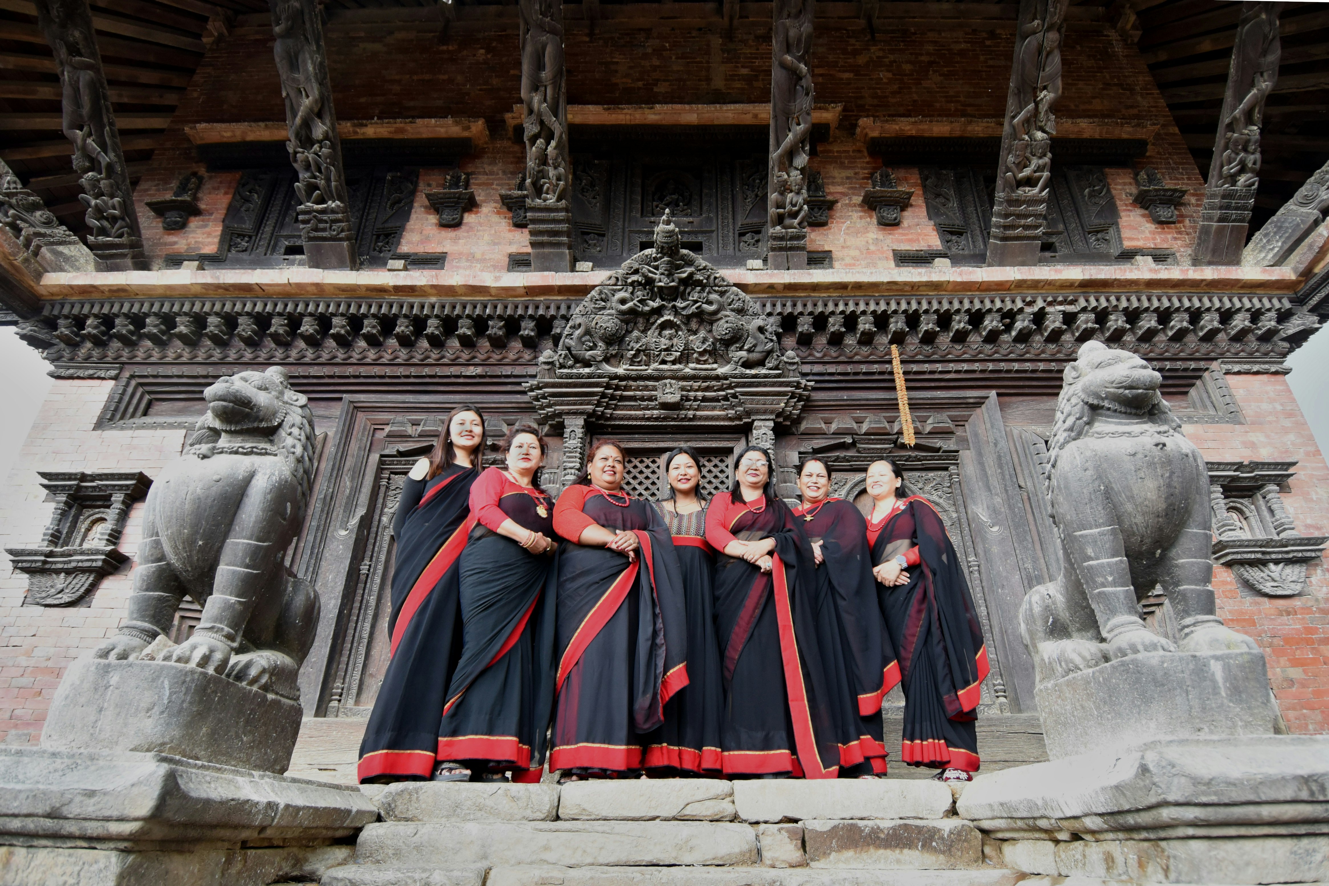 A group of women, all wearing a full-length black and red gown, stand in front of an ornately carved wooden door