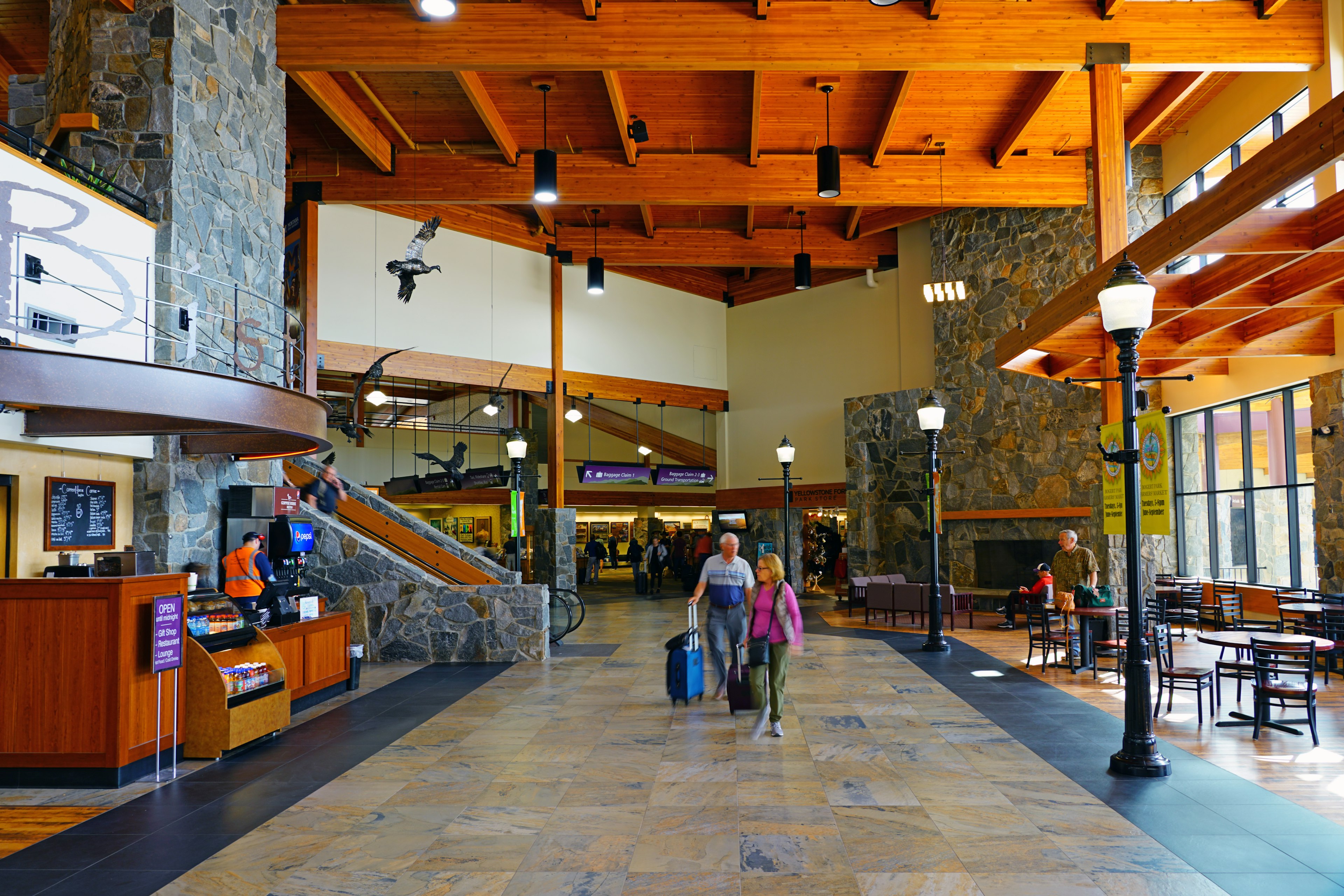 A couple wheel luggage through a quiet airport with large windows and wooden beams