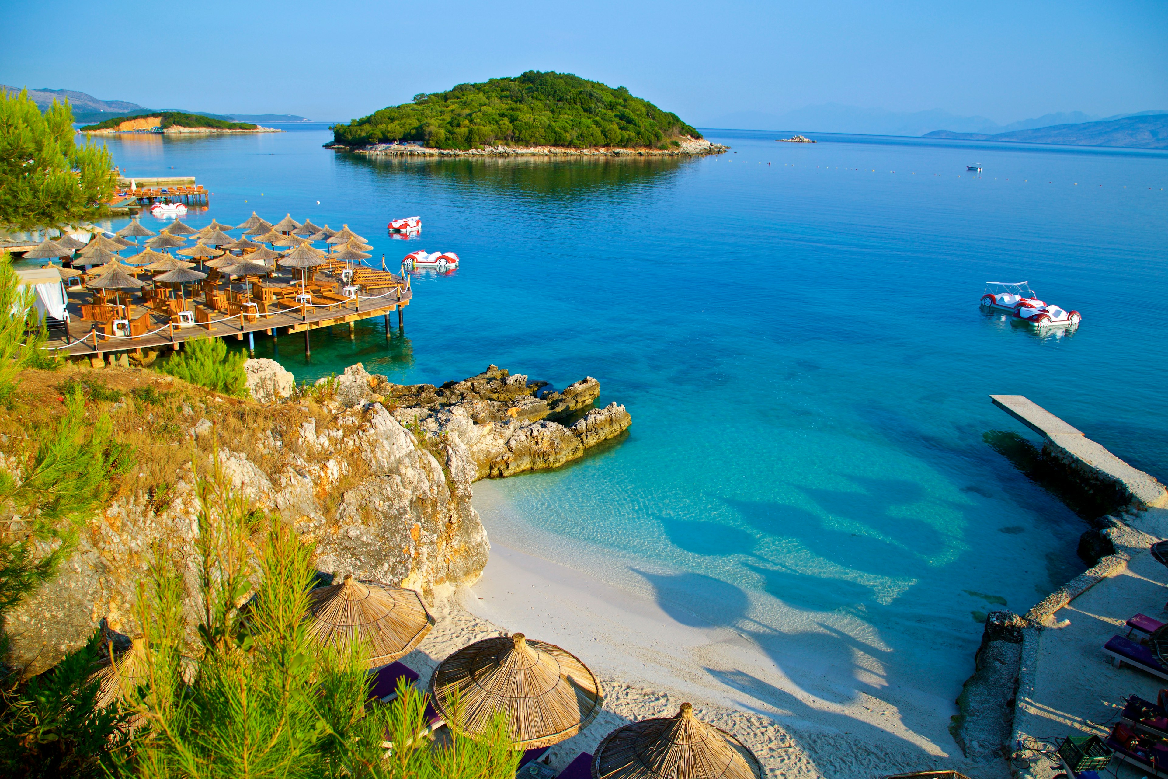 A wooden pier on the right, a collection of tables on the left, and a beach in the middle stretches into shallow blue waters off the coast of Ksamil beach, with a small island, covered in trees, in the distance.