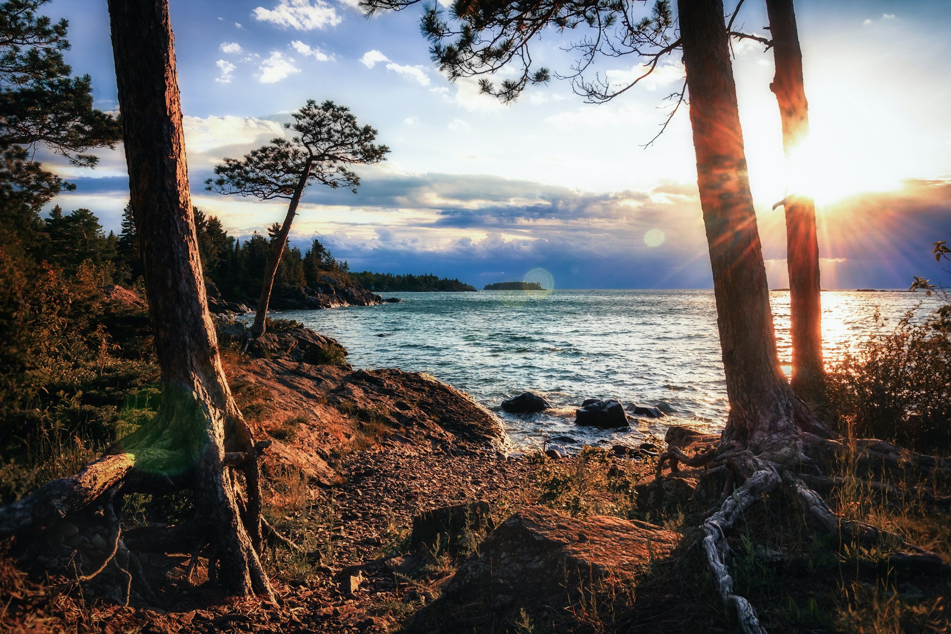 A rocky shoreline of a lake viewed from woodland as the sun sets