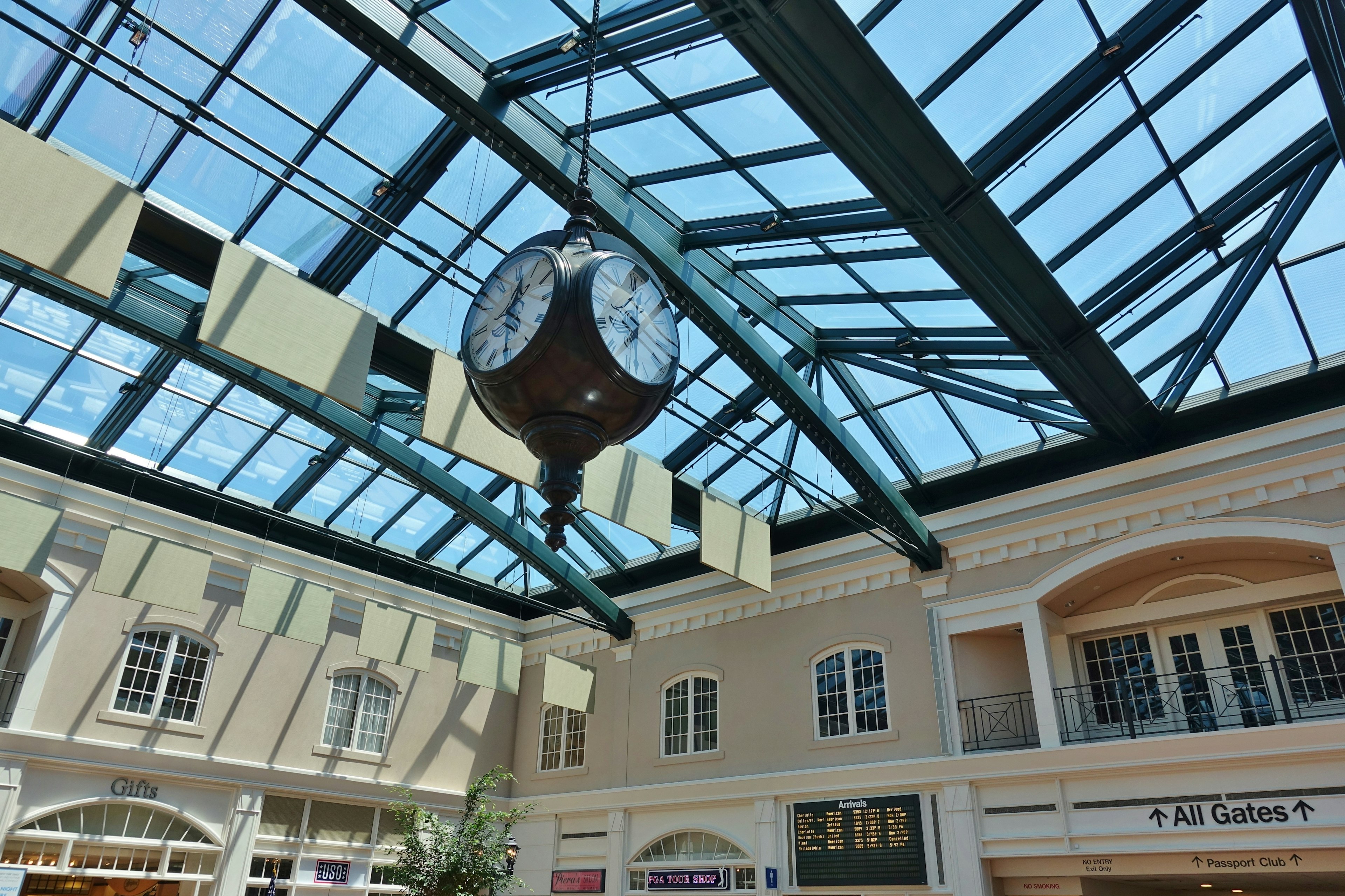 A large clock hangs from a glass ceiling in an atrium designed like a town square