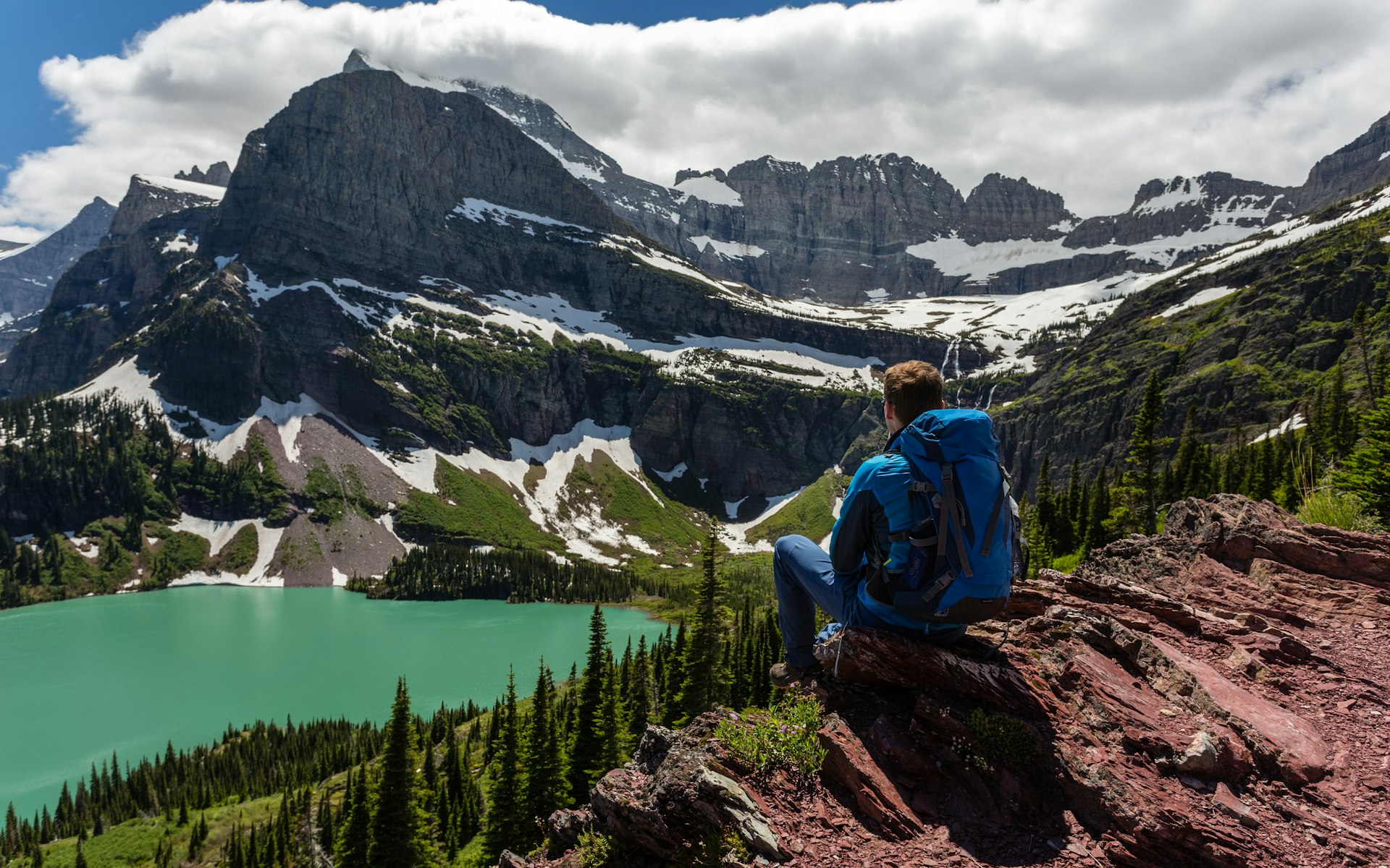 A hiker sits on a precipice looking down over an alpine lake