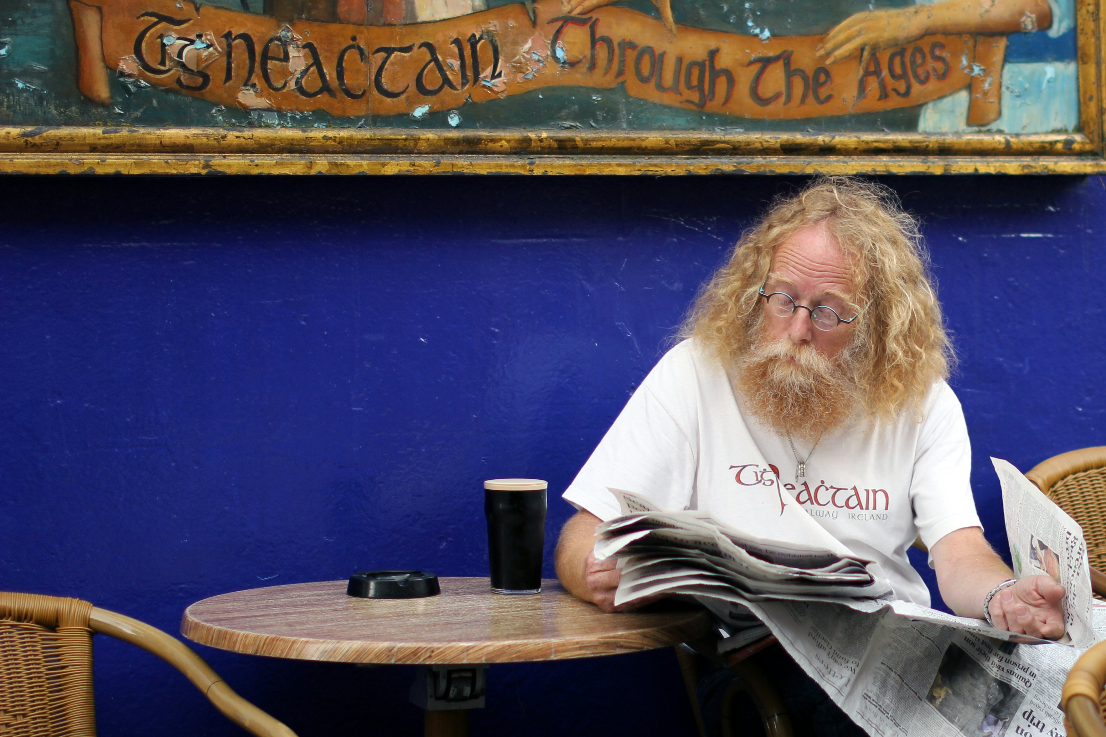 A man with long hair drinks a pint of Guinness outside a blue pub.