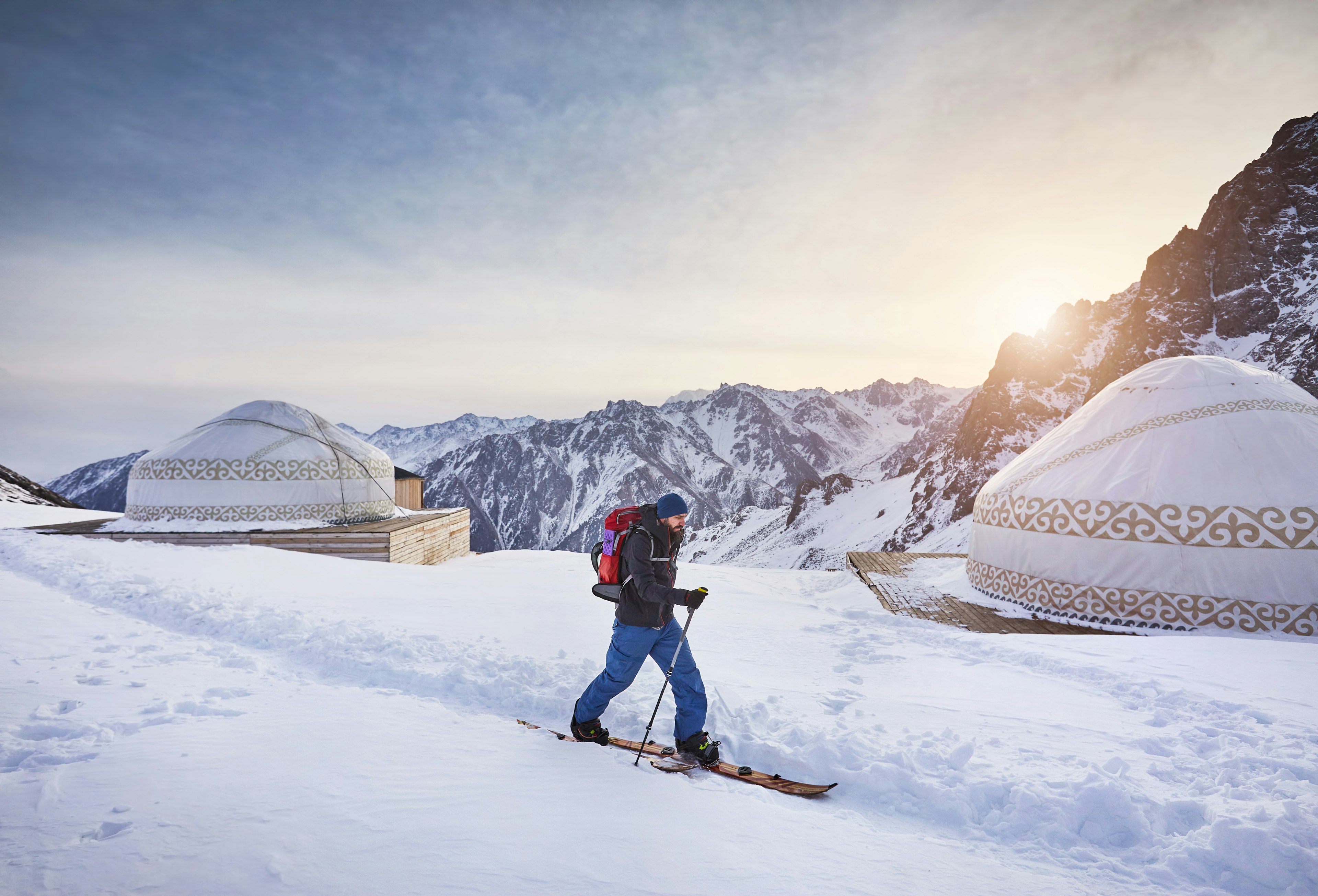 A man on skis ski touring uphill with beard and backpack on a high snowy mountain near traditional nomad yurt house at ski resort Shymbulak in Almaty, Kazakhstan