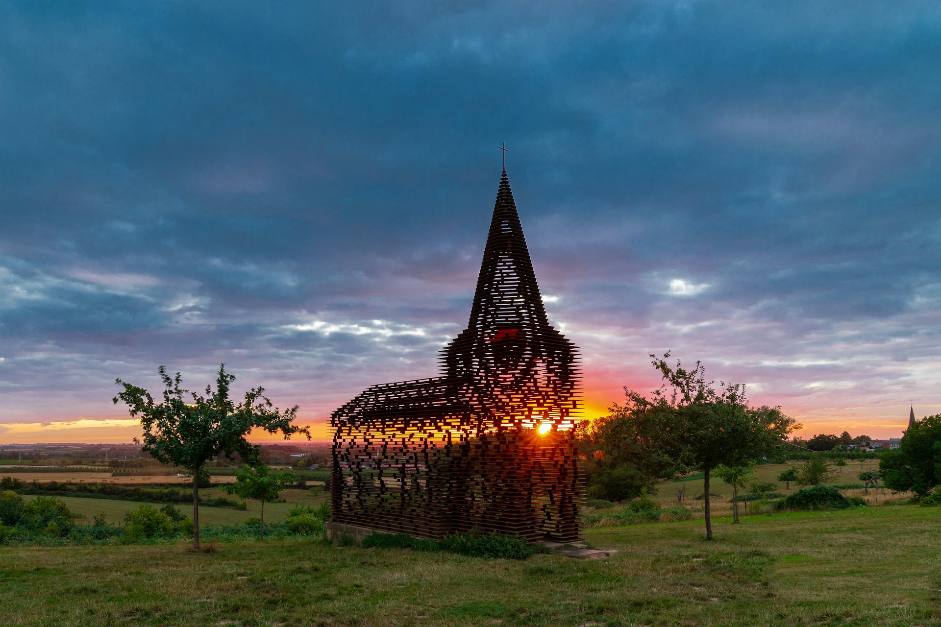 Uma escultura feita de ripas em forma de estrutura semelhante a uma capela, com o sol poente brilhando através dela
