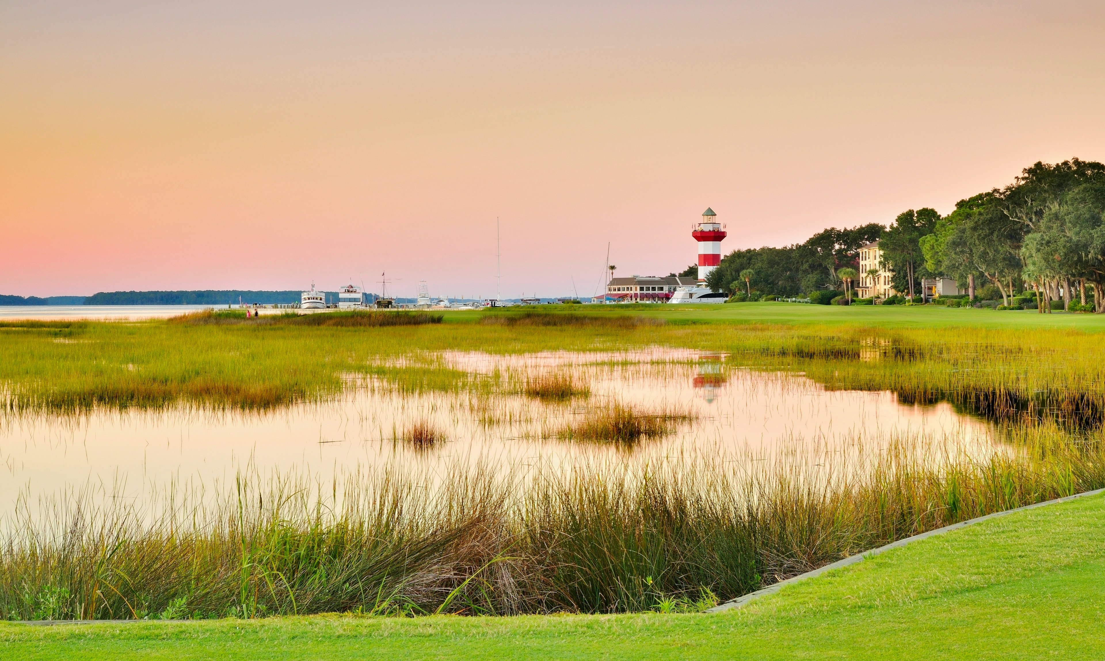 The edge of a golf green right near the water and reeds that line the harbor. A red-and-white lighthouse stands in the distance