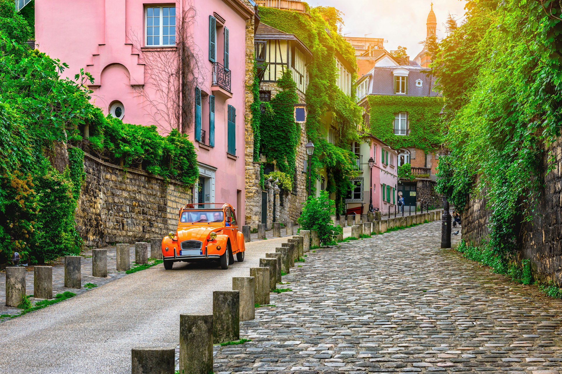 An orange Citroën 2CV car travels down an old street in the Montmartre quarter of Paris