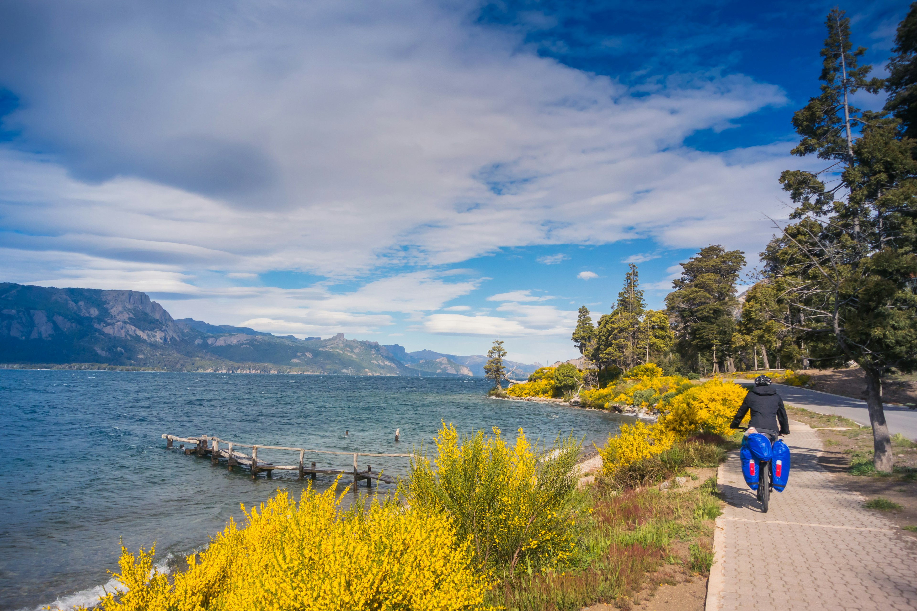 A cyclist pedals along on a bike with bags on the back beside a lake in a mountainous area