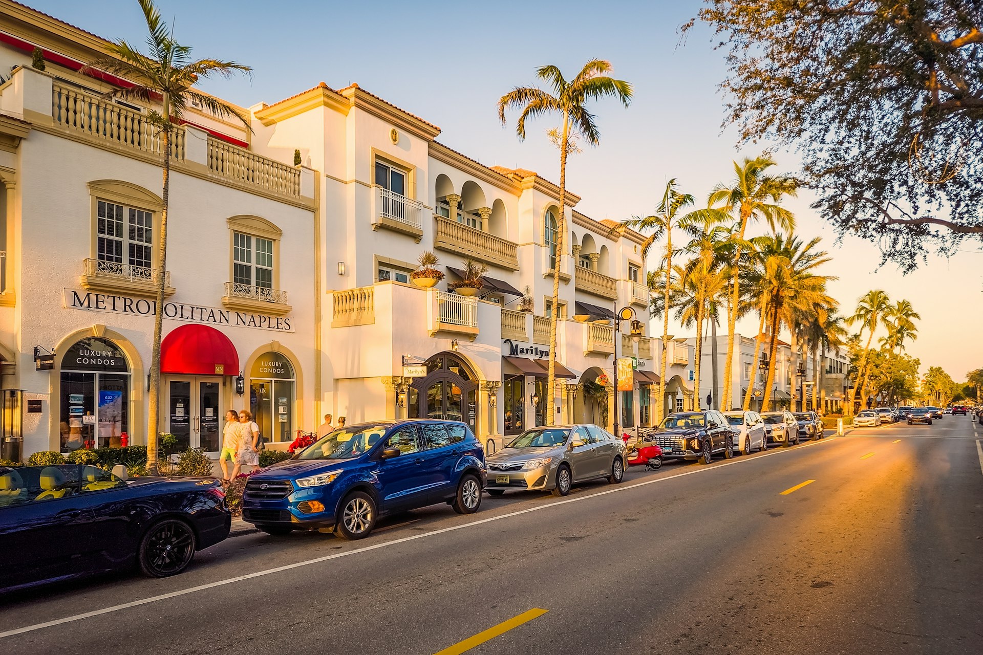 Cars are parked alongside white buildings on a street lined with palm trees at sunset