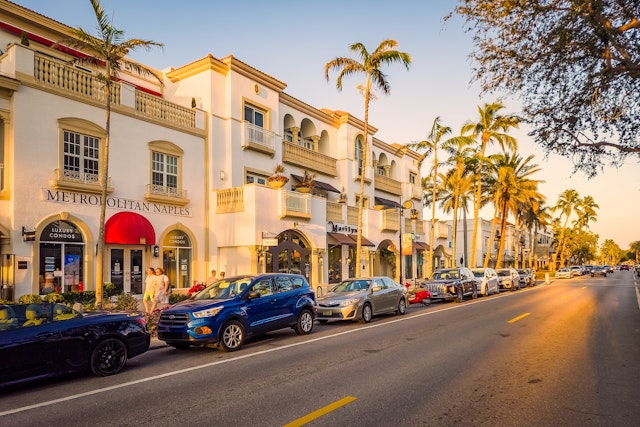 Cars are parked alongside white buildings on a street lined with palm trees at sunset