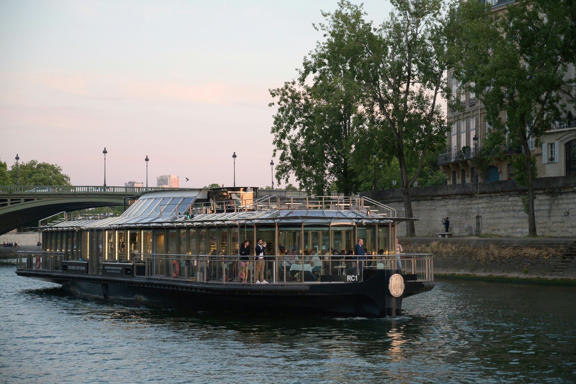 Some people are standing out on the deck of the Ducasse sur Seine, a floating restaurant, as it glides down the River Seine at dusk.