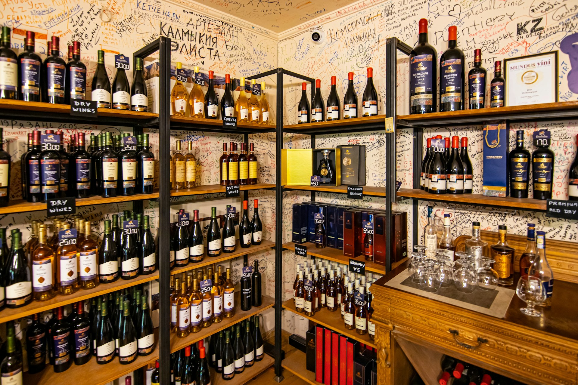 Uma loja de vinhos tem prateleiras repletas de diferentes variedades de vinho georgiano;  as paredes atrás das prateleiras são cobertas por letras em estilo graffiti.
