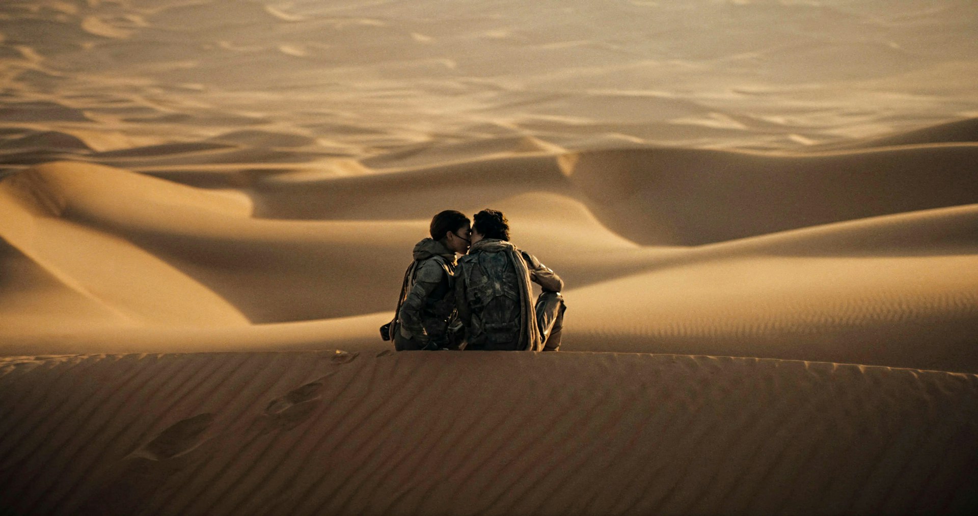 Two people sitting on sand dunes in a vast desert lean in to kiss